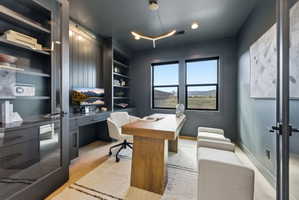 Dedicated office space featuring two large windows, built-in shelving, a full-wall desk unit, and light wood-finish flooring