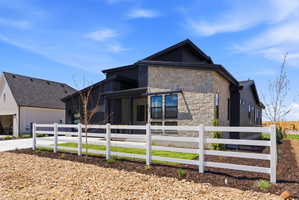 Modern architectural design featuring stone and dark siding, multiple large windows, a covered entry, and a white fence