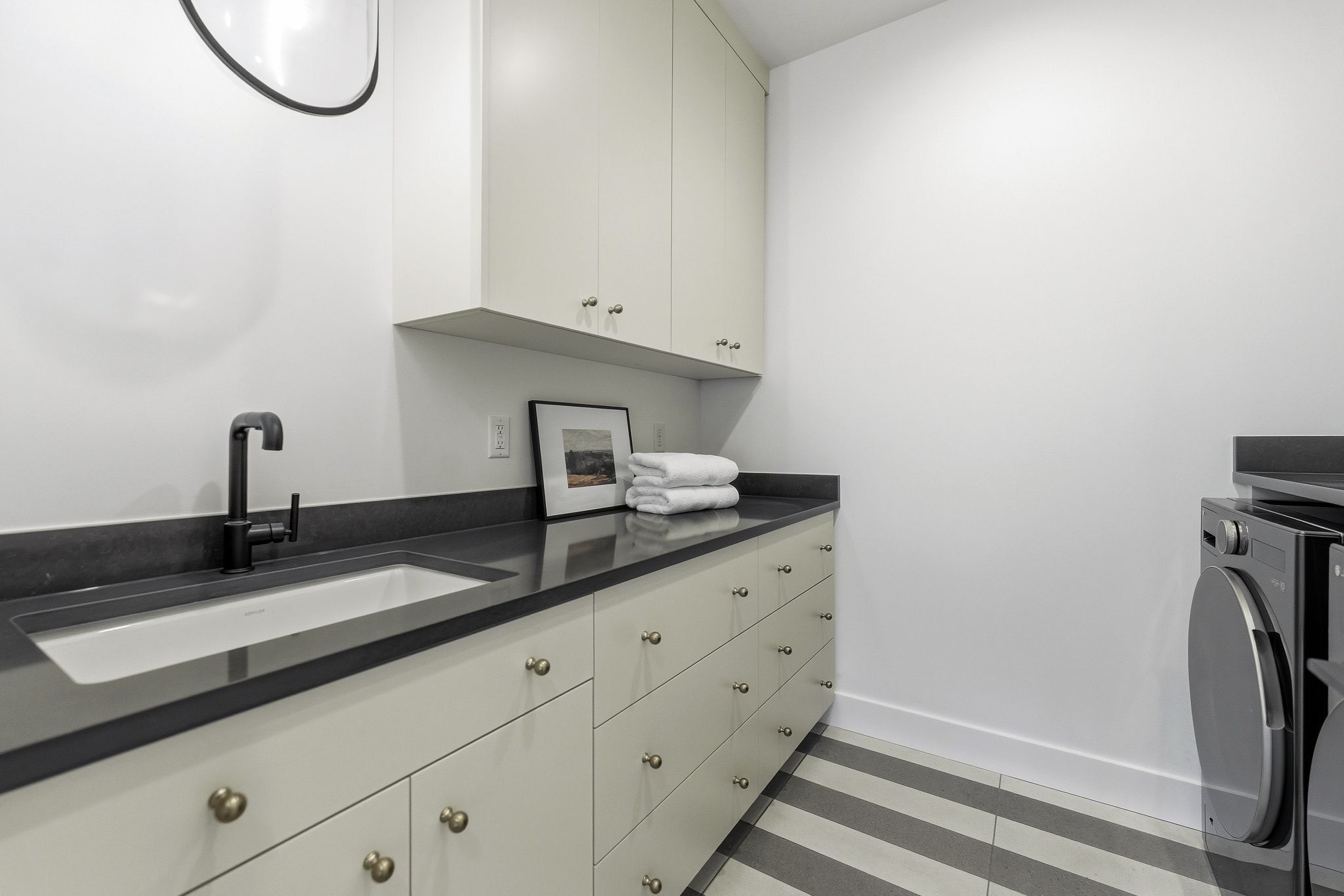 Utility room featuring light-colored cabinetry with brass hardware, a dark countertop, an undermount sink with a matte black faucet, and striped tile flooring