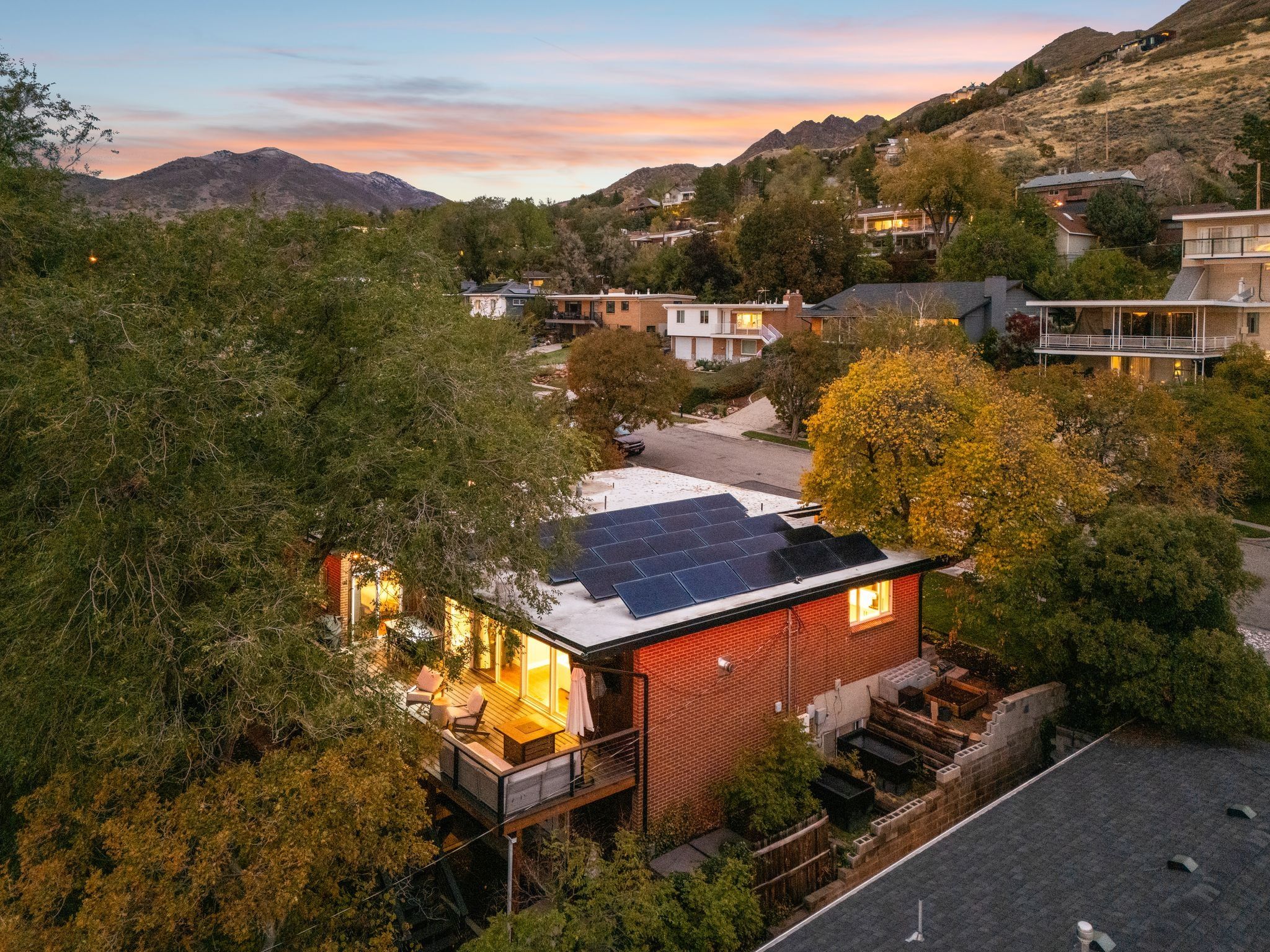 Red brick exterior featuring a multi-panel solar array, expansive upper-level deck, surrounding mature trees, and tiered landscaping