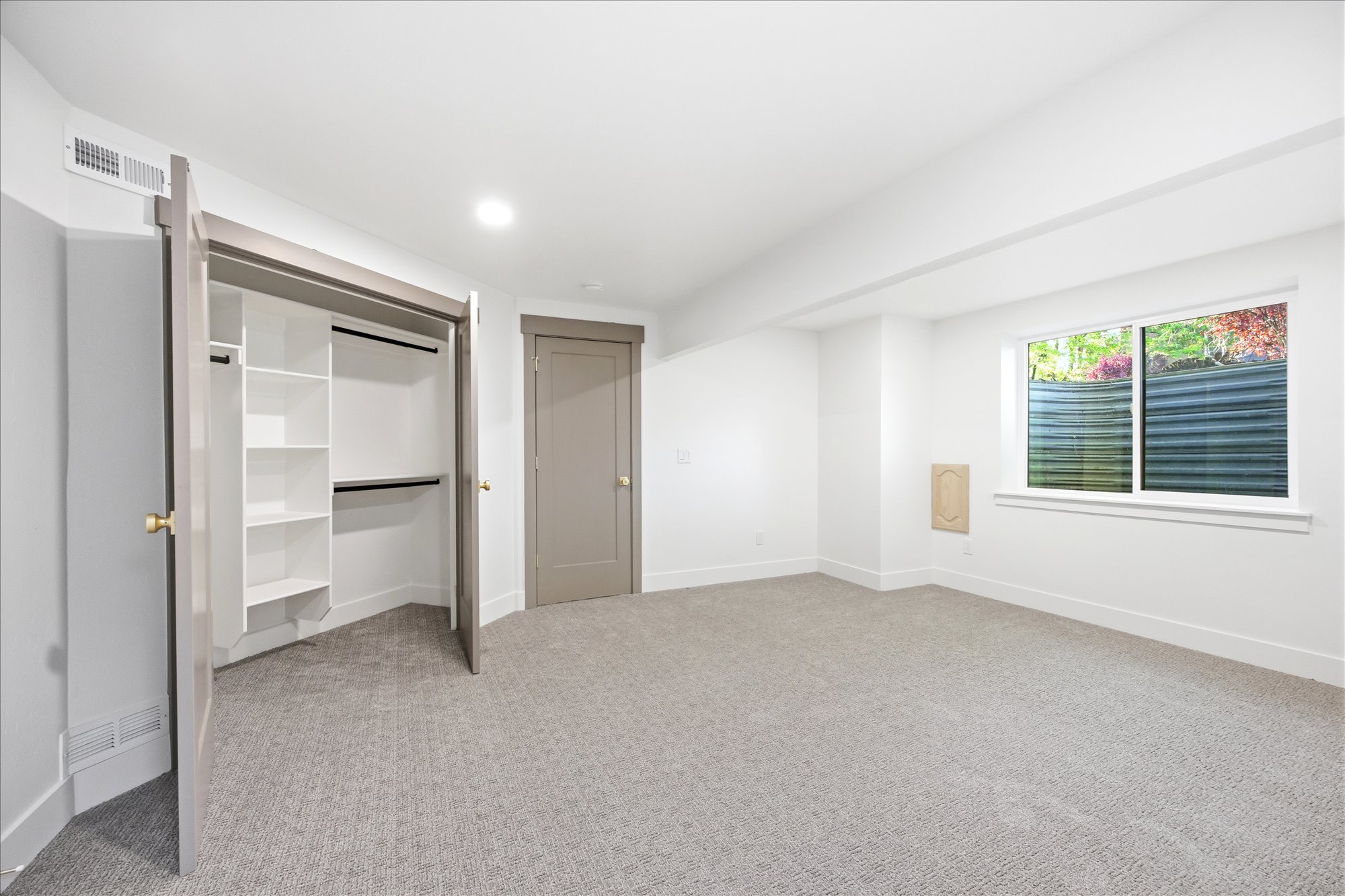 Spacious room with light gray patterned carpeting, white walls, and a large window with exterior foliage