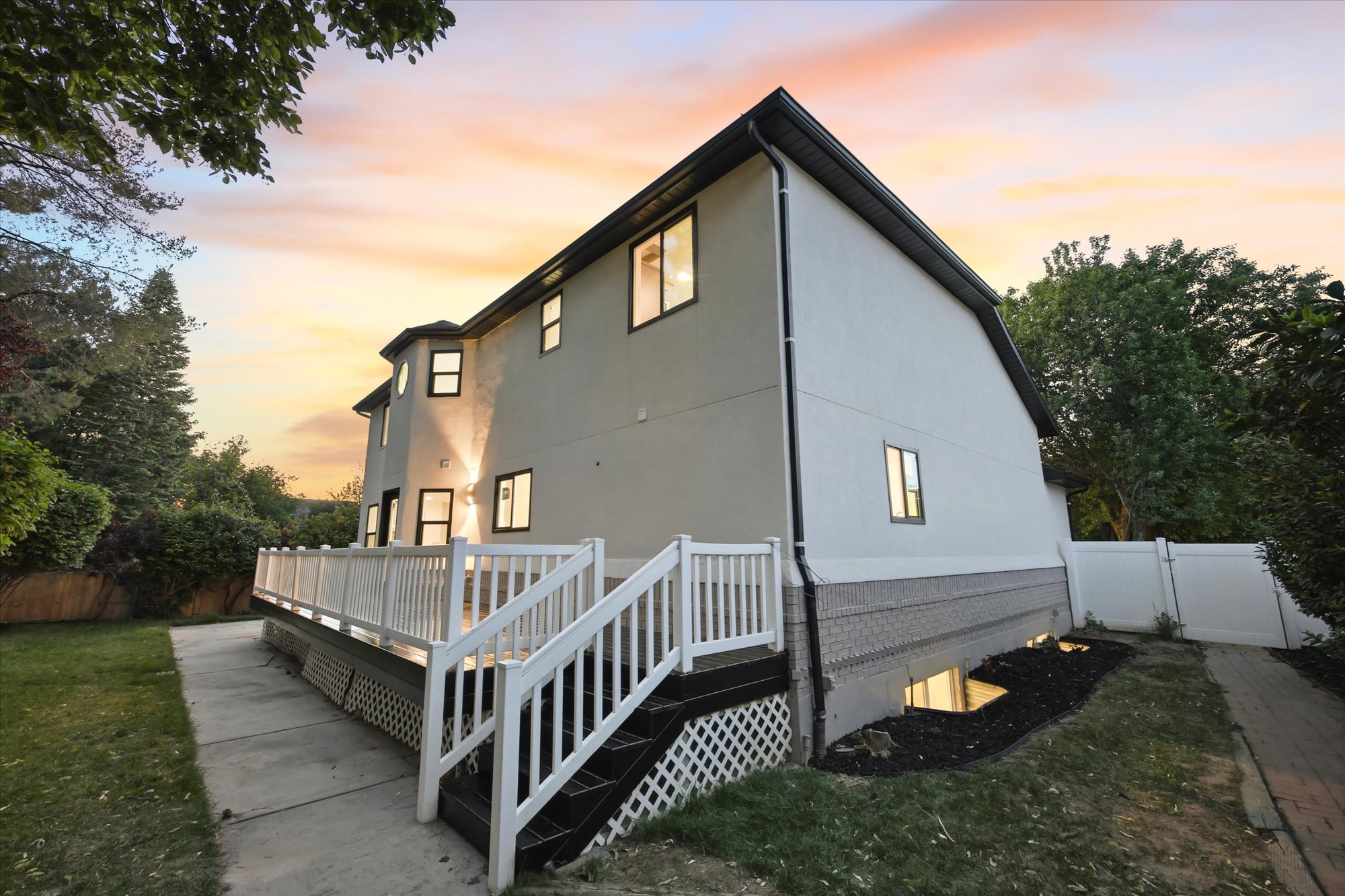 Rear exterior showcasing a white railing deck with steps, a stucco finish, multiple windows, and a foundation with grey brick detailing