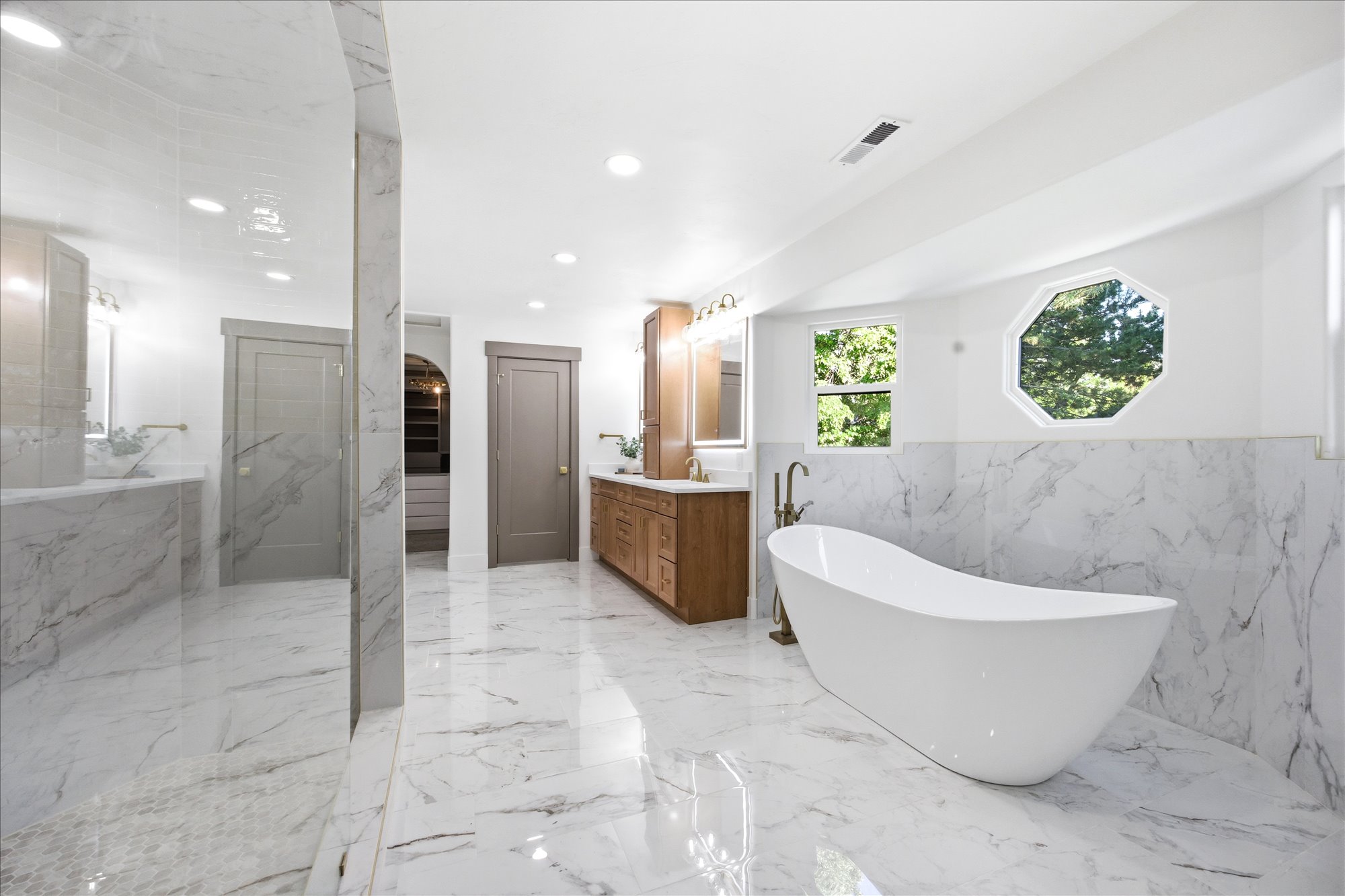 Spacious bathroom featuring a white freestanding soaking tub with floor-mounted faucet, octagonal window, and white marble-look tile flooring