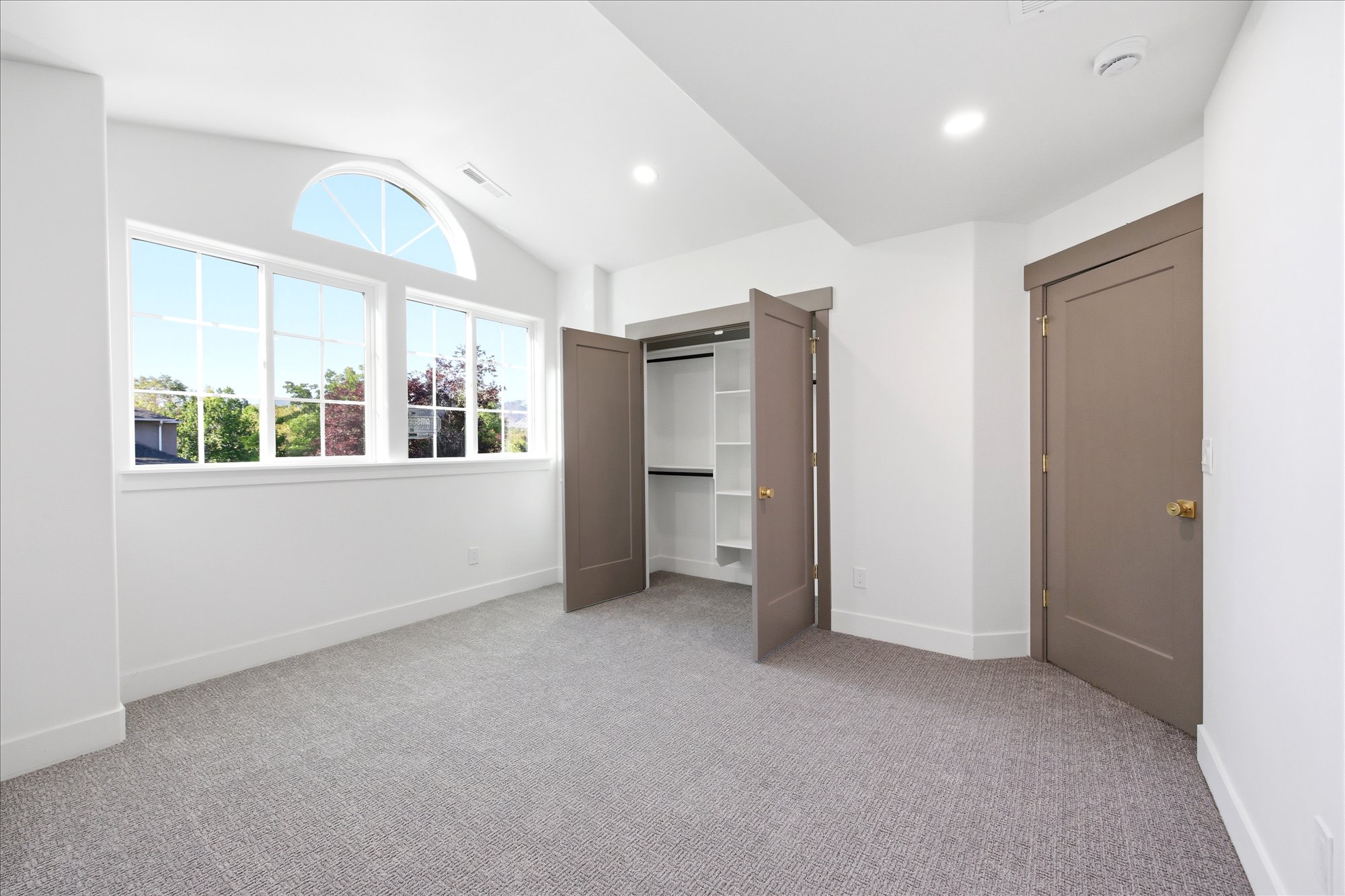 Room featuring a large window array with an arched transom, recessed ceiling lighting, textured gray carpeting, a built-in closet with shelving, and two panel doors with brass hardware