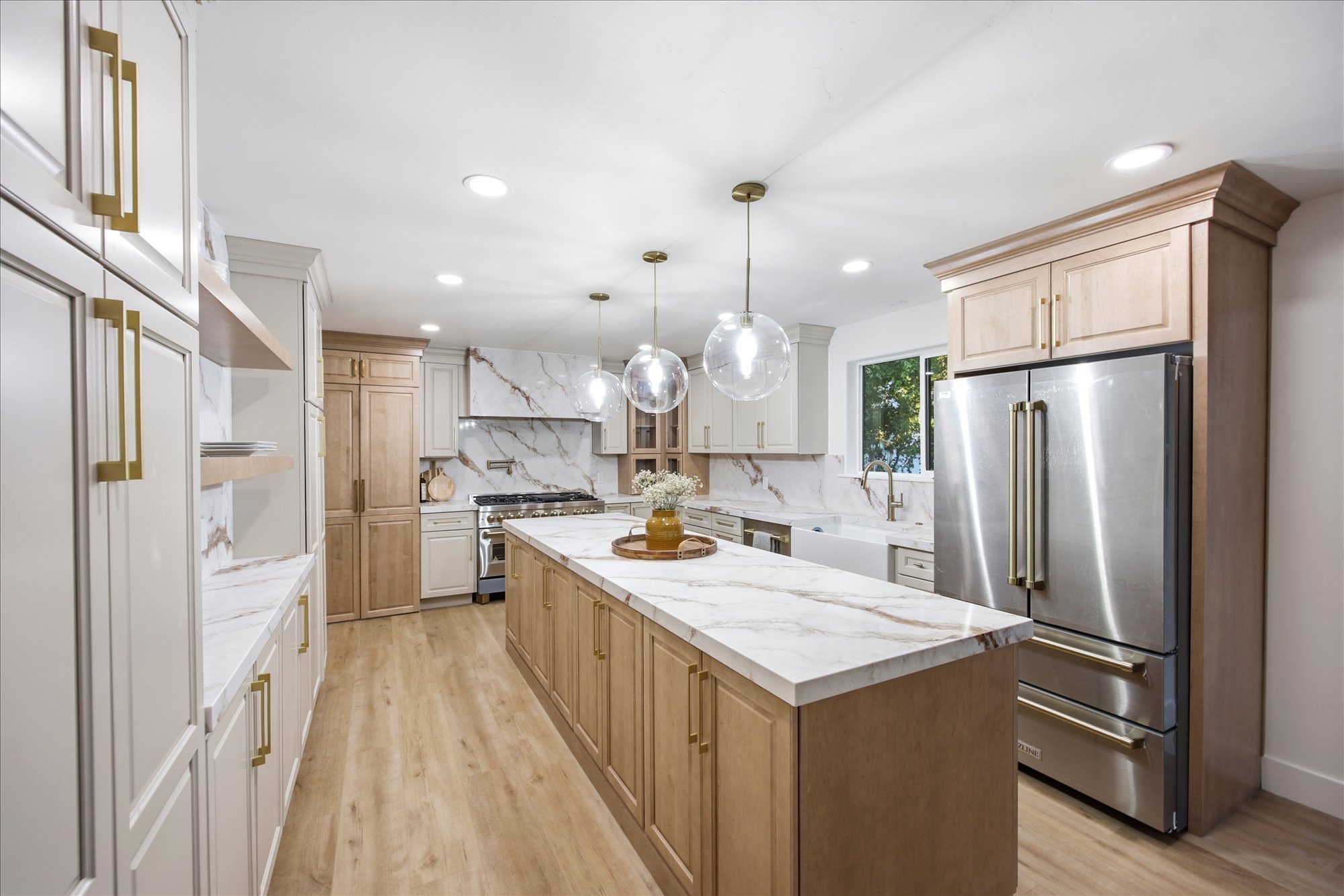 Spacious kitchen featuring a large central island with a white veined countertop, stainless steel appliances, light wood-finish flooring, and white cabinetry with gold-tone hardware