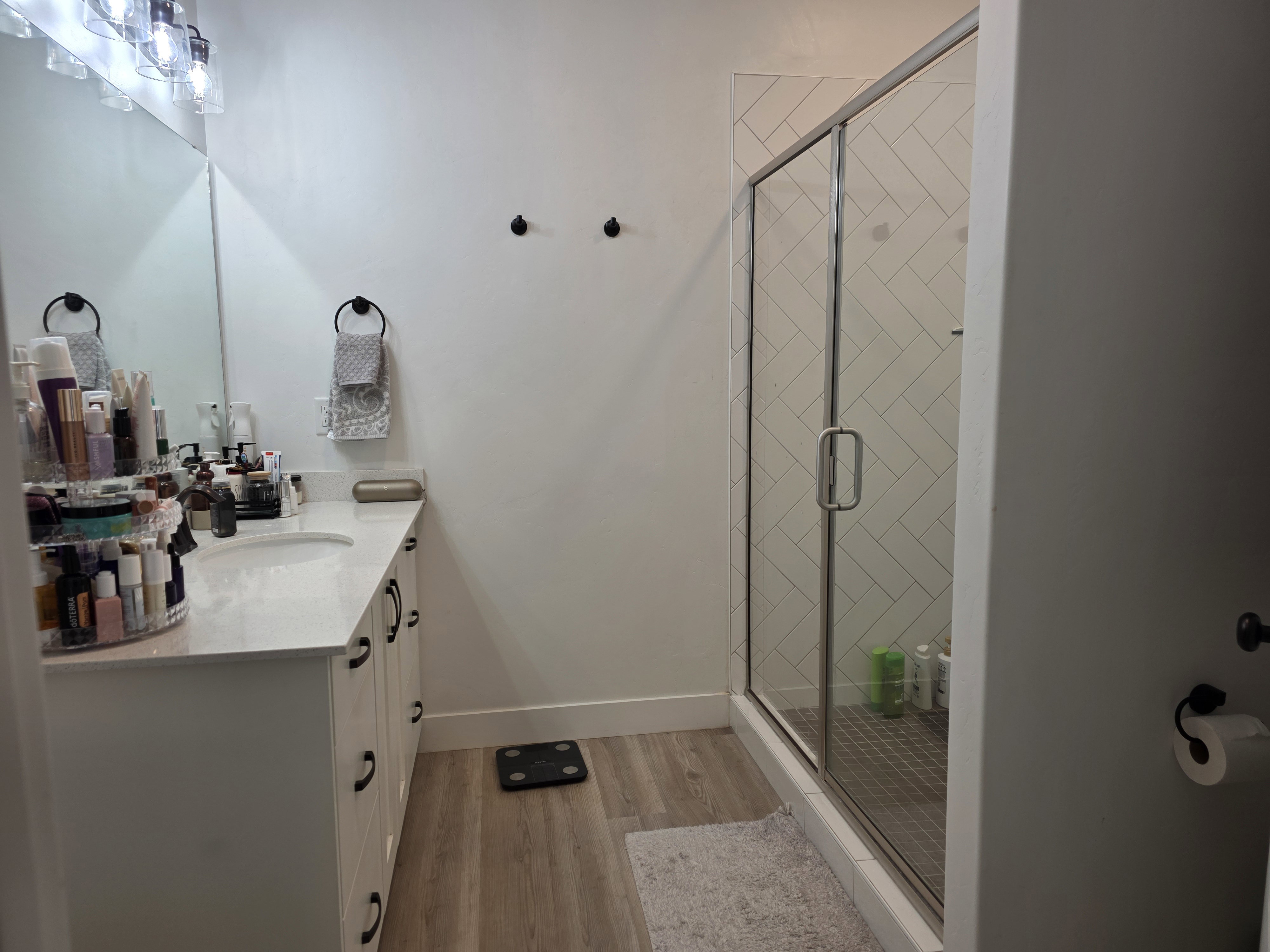 Bathroom featuring a single vanity with a white countertop and undermount sink, wood-finish flooring, a glass-enclosed shower with subway tile surround, and contemporary black hardware