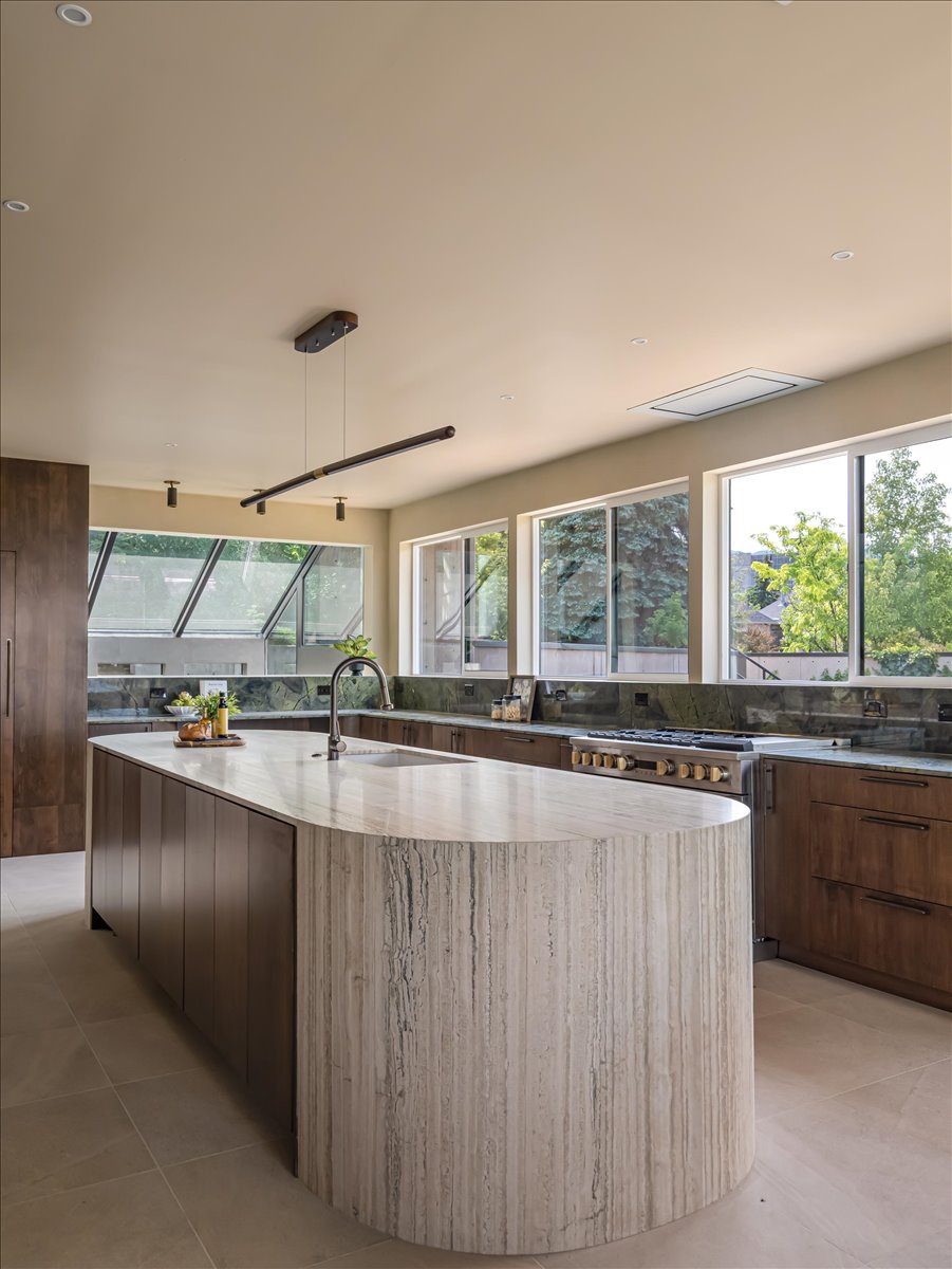 Contemporary kitchen featuring a substantial stone island with integrated sink, wood-finish cabinetry, a professional-grade range, and a full wall of picture windows