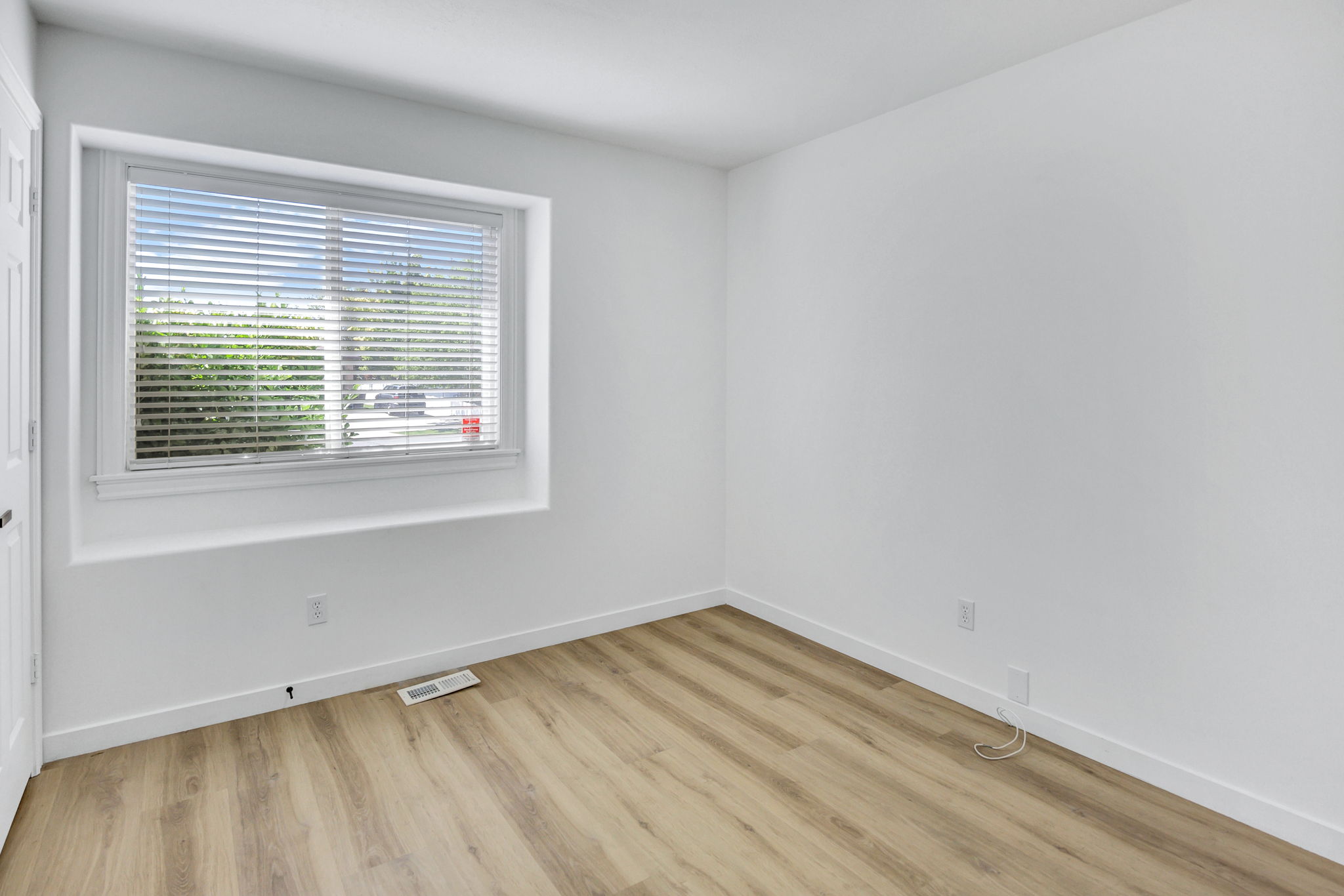 Bright room featuring light wood-finish flooring, crisp white walls, and a large window with white blinds