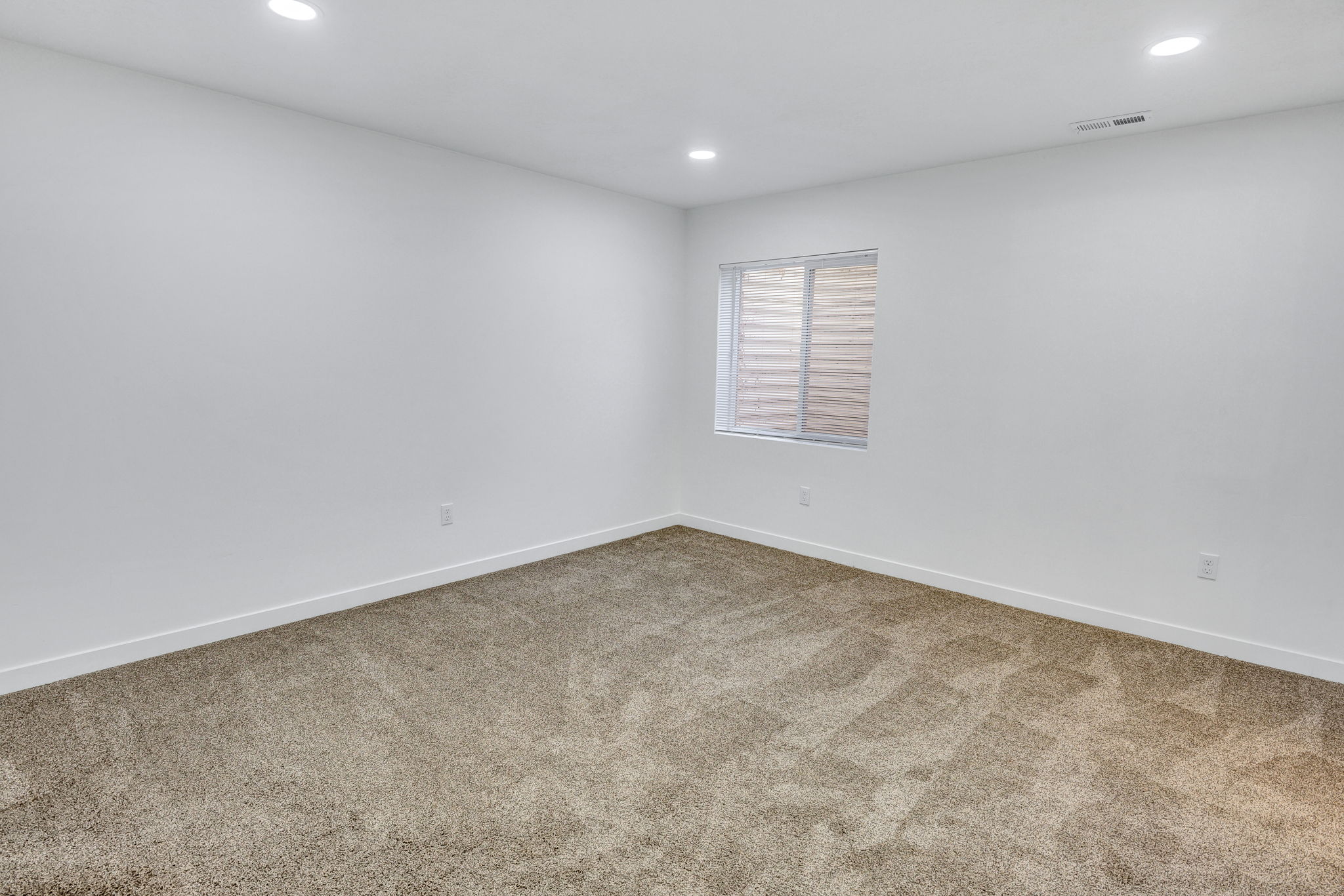 Spacious room featuring neutral carpeting, white walls, recessed ceiling lighting, and a window with horizontal blinds