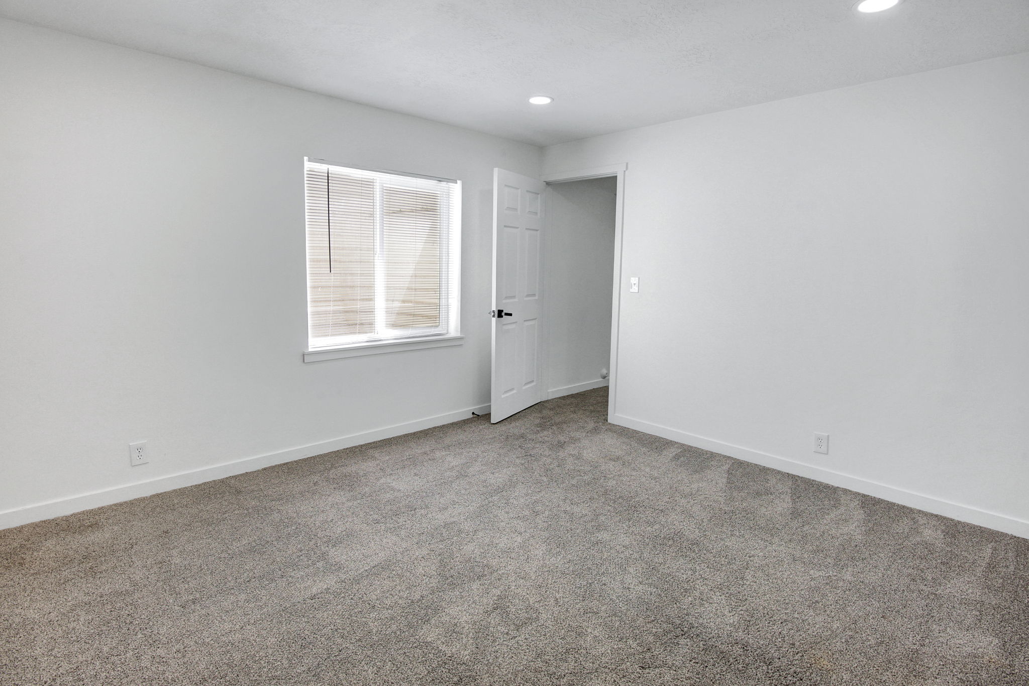 Spacious interior room featuring light gray carpeting, white walls, recessed ceiling lighting, a window with horizontal blinds, and a white interior door with a black handle
