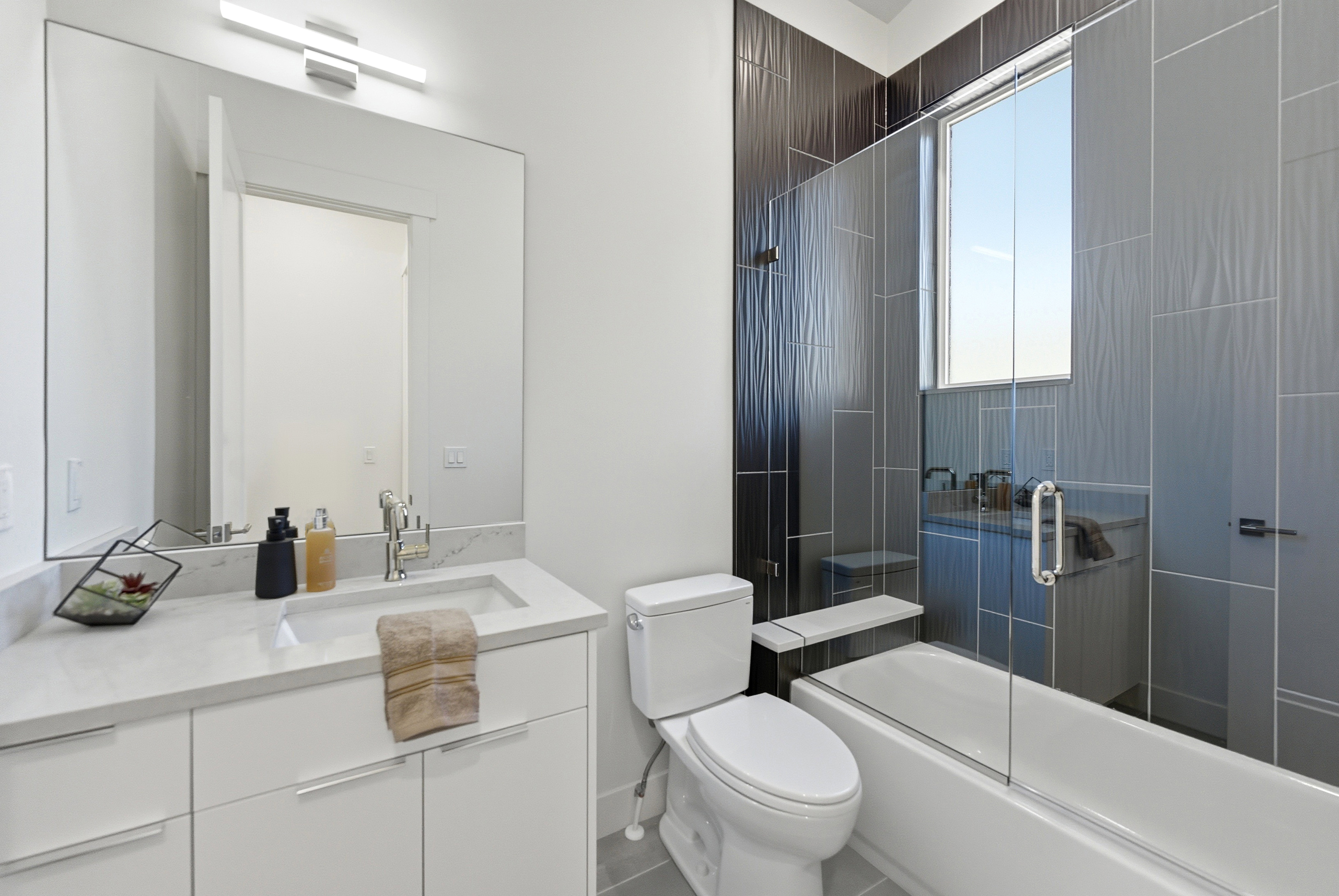 Contemporary bathroom featuring a white vanity with an undermount sink and a polished chrome faucet, a large rectangular mirror with an overhead linear light fixture, a toilet, and a tub/shower combination with a clear glass enclosure and textured dark gr