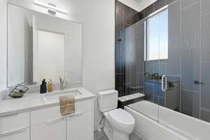 Contemporary bathroom featuring a white vanity with an undermount sink and a polished chrome faucet, a large rectangular mirror with an overhead linear light fixture, a toilet, and a tub/shower combination with a clear glass enclosure and textured dark gr