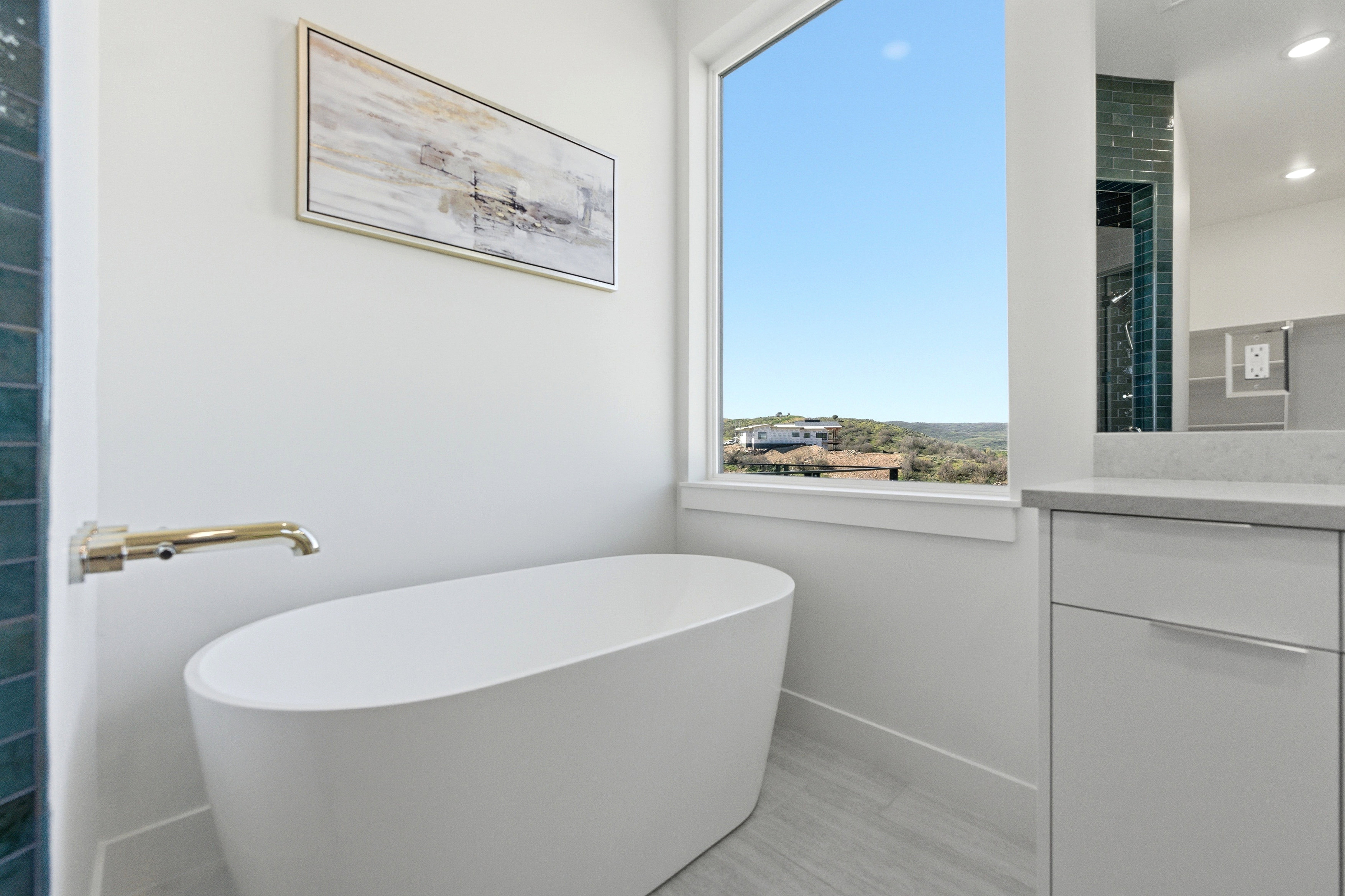 Primary bath features freestanding oval soaking tub with a wall-mounted brass faucet, large window, tile flooring, and a vanity featuring a light-toned countertop