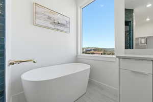 Primary bath features freestanding oval soaking tub with a wall-mounted brass faucet, large window, tile flooring, and a vanity featuring a light-toned countertop
