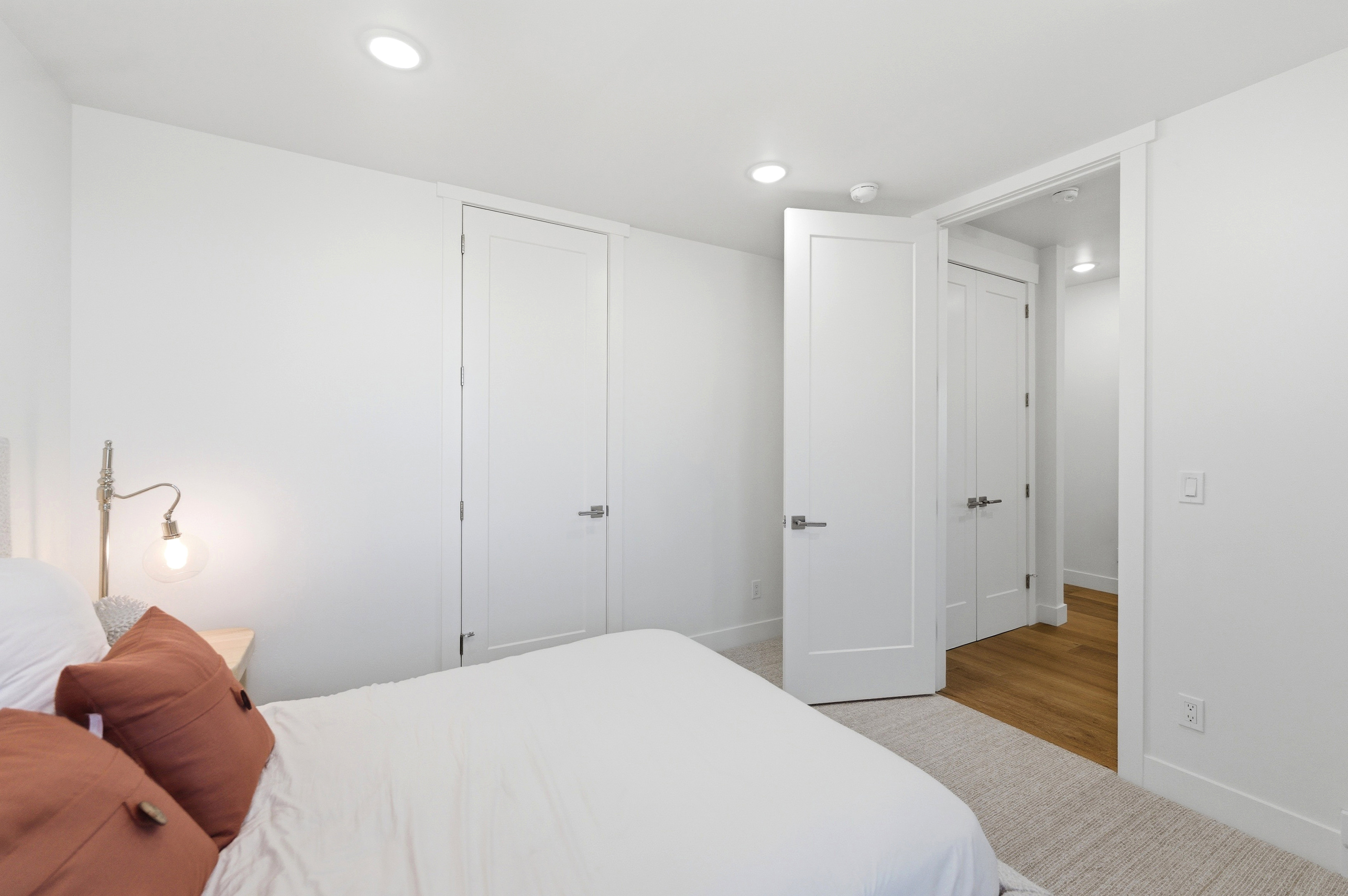 Recessed ceiling lighting illuminates a room featuring a neutral color palette, multiple white paneled doors, and a light-toned carpet leading to wood-finish flooring in the entryway