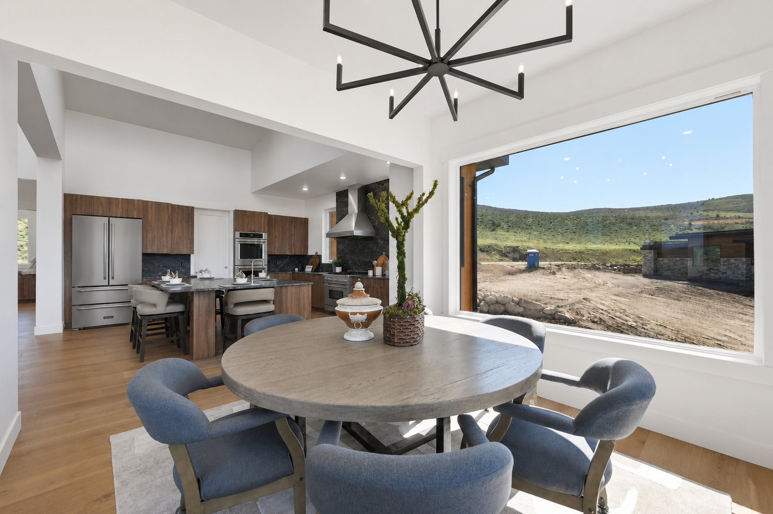 Open-concept kitchen and dining area featuring a prominent window, stainless steel appliances, dark wood-finish cabinetry, light wood-finish flooring, and a contemporary chandelier