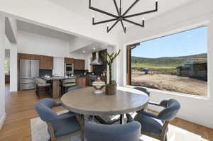 Open-concept kitchen and dining area featuring a prominent window, stainless steel appliances, dark wood-finish cabinetry, light wood-finish flooring, and a contemporary chandelier