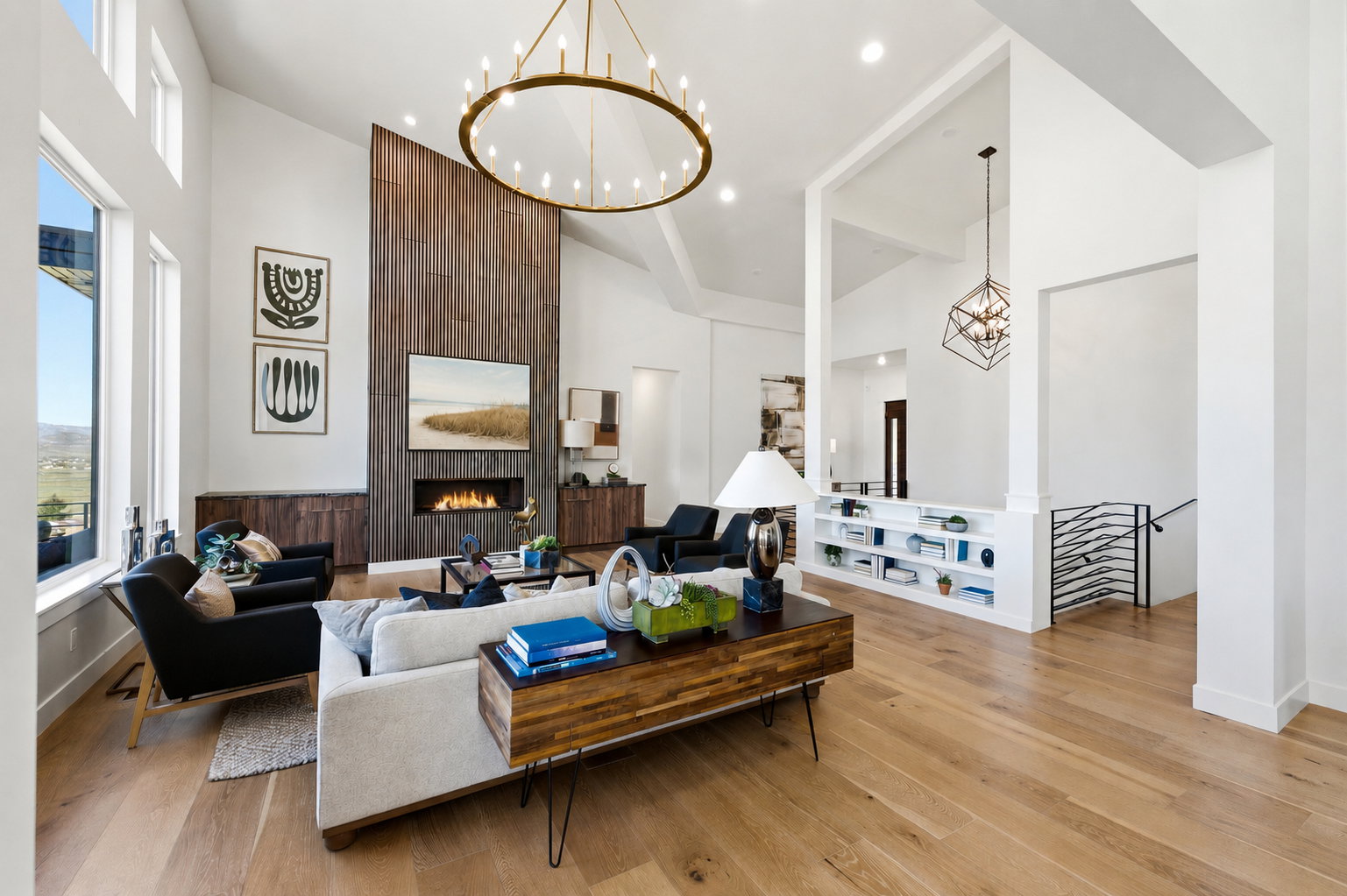 Spacious living area featuring high vaulted ceilings, expansive windows, light wood-finish flooring, a contemporary fireplace with a wood-paneled accent wall, and a large circular chandelier, TV virtually staged