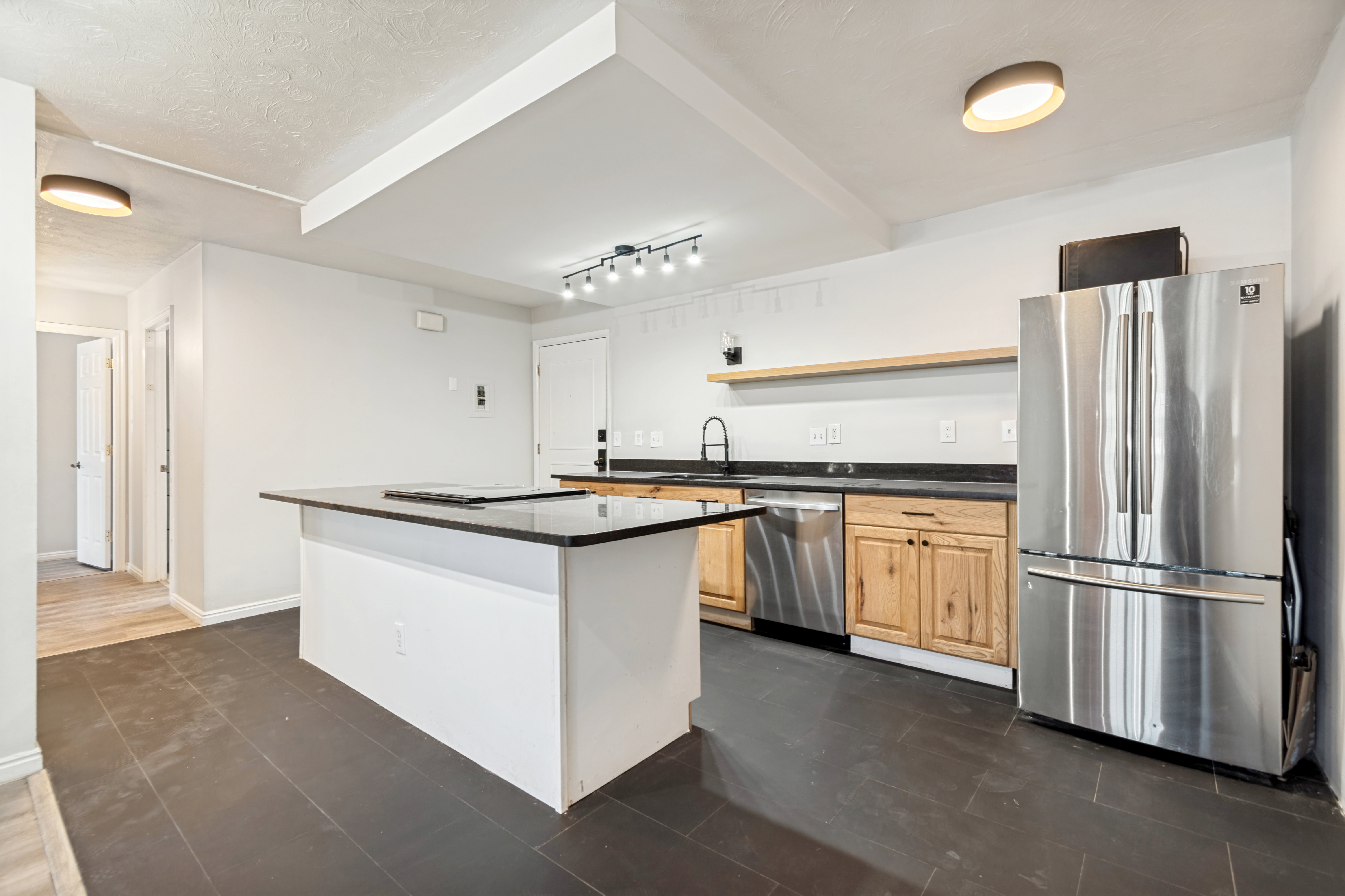 Open-concept kitchen featuring dark tile flooring, a center island with a dark countertop, light wood cabinetry, stainless steel appliances, and a floating wood shelf