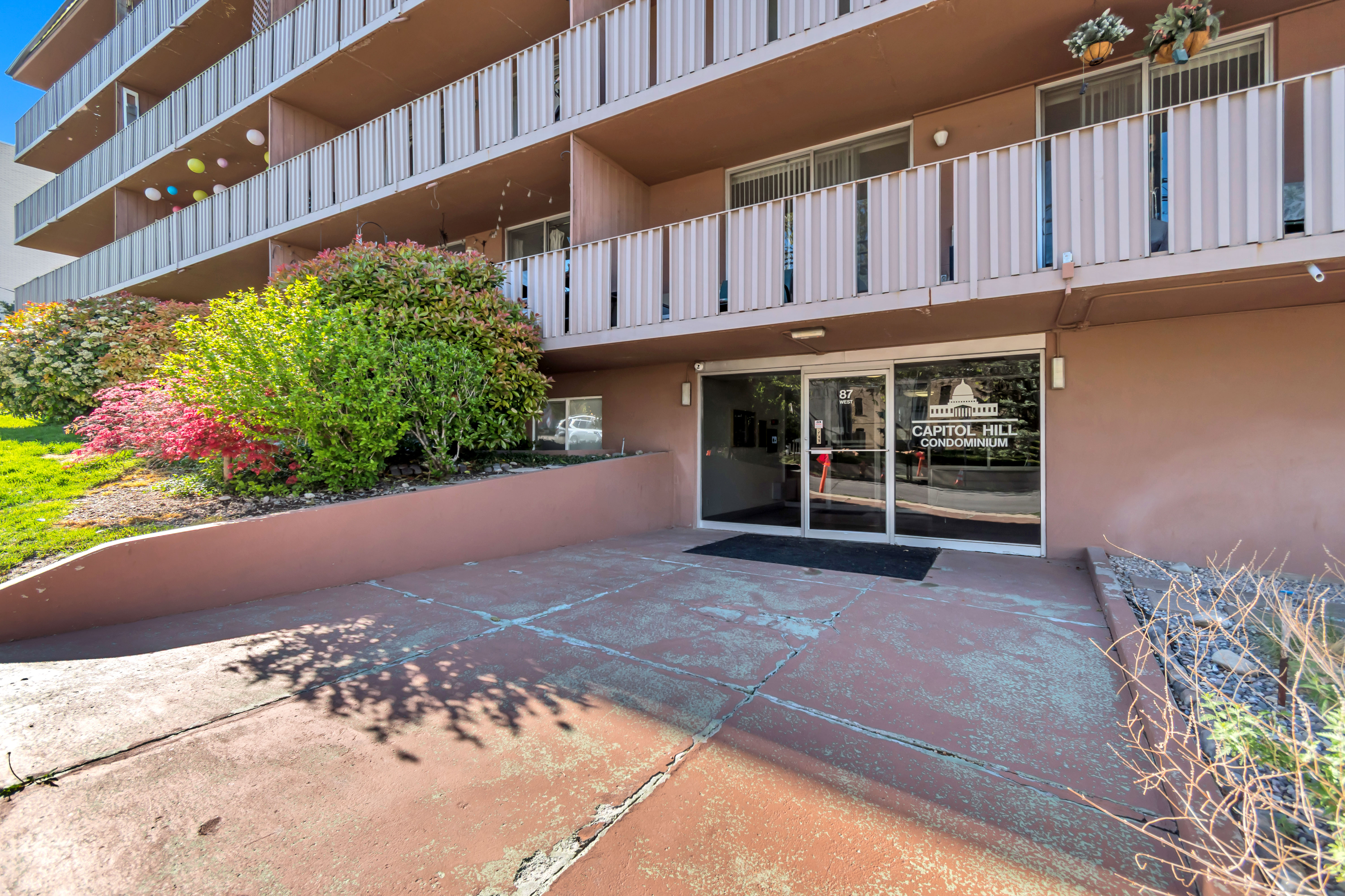 Capitol Hill Condominium building entrance featuring a glass door, a dark entryway mat, and an adjacent landscaped area with mature plantings