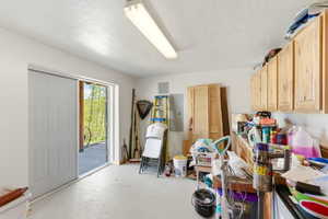 Utility room featuring painted concrete flooring, overhead fluorescent lighting, a sliding white panel door, and light wood cabinetry with upper storage
