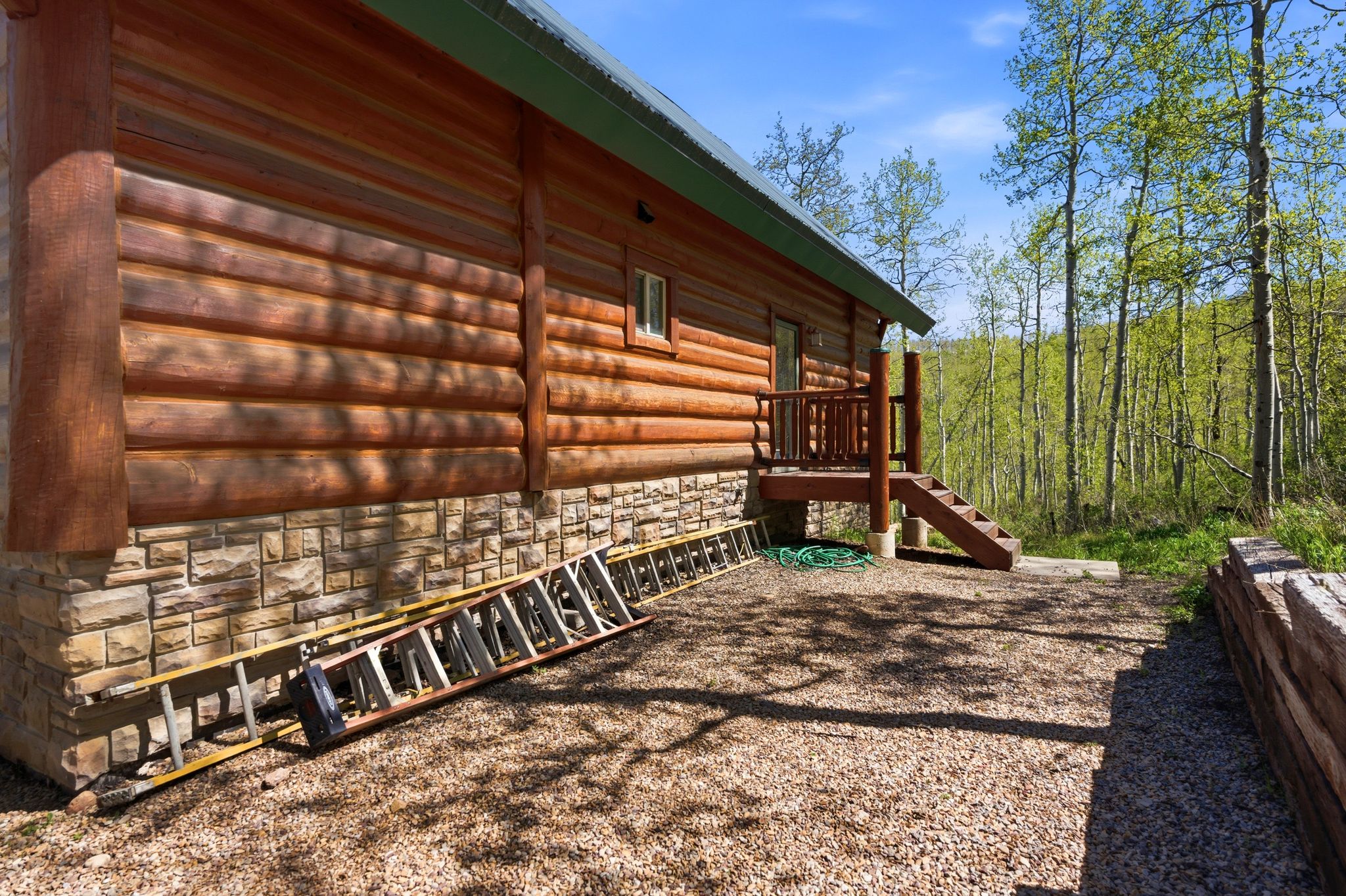 Wood-sided log cabin with a stone foundation, featuring a wood deck and stairs, set against a backdrop of mature trees