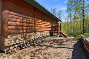 Wood-sided log cabin with a stone foundation, featuring a wood deck and stairs, set against a backdrop of mature trees