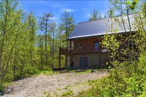 Wood-sided cabin-style residence featuring a metal roof, a second-story balcony with wood railing, a stone-faced lower level, and surrounding mature trees