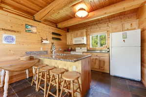 Kitchen featuring wood-paneled walls and ceiling, exposed wood beams, slate-style floor tiling, light wood cabinetry, and stone-finish countertops