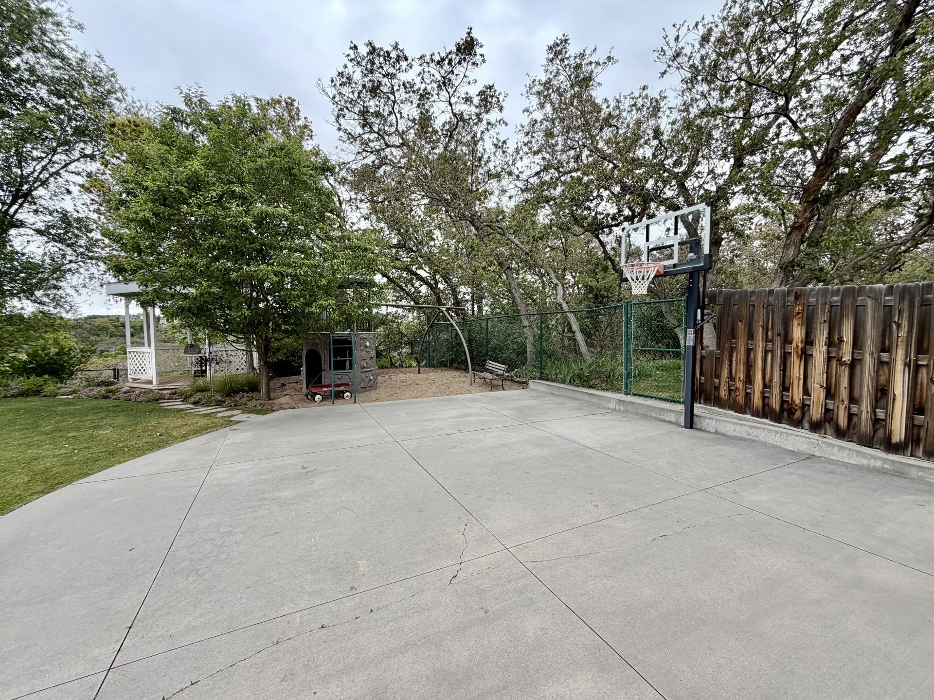 Spacious concrete patio featuring a basketball hoop, surrounded by mature trees and a wooden privacy fence