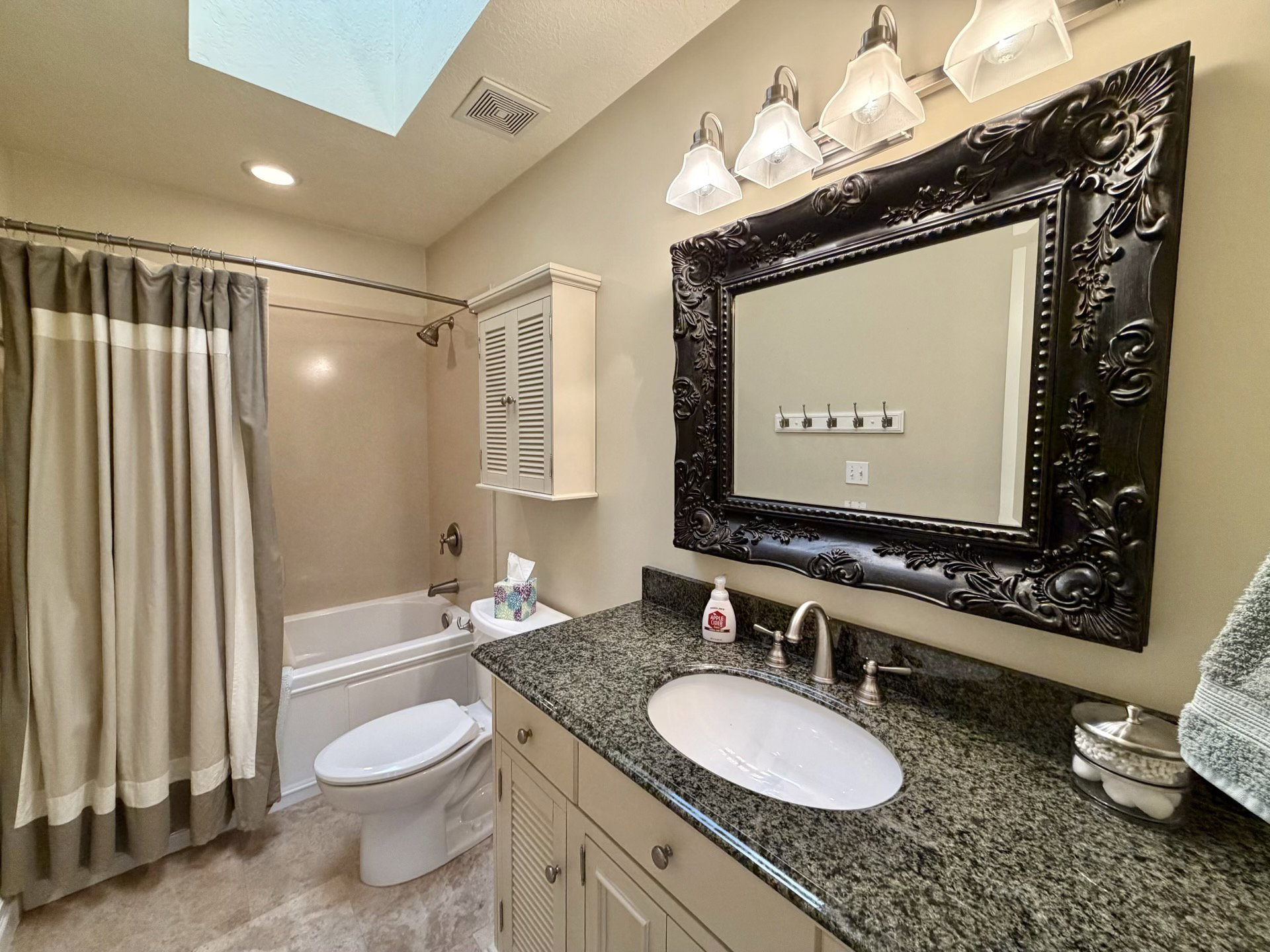 Bathroom featuring a skylight, a single vanity with a speckled countertop, an ornate framed mirror, and a built-in bathtub with shower