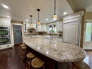 Spacious kitchen featuring a large granite-topped island, double wall ovens, paneled refrigerator, farmhouse sink, and rich wood-finish flooring