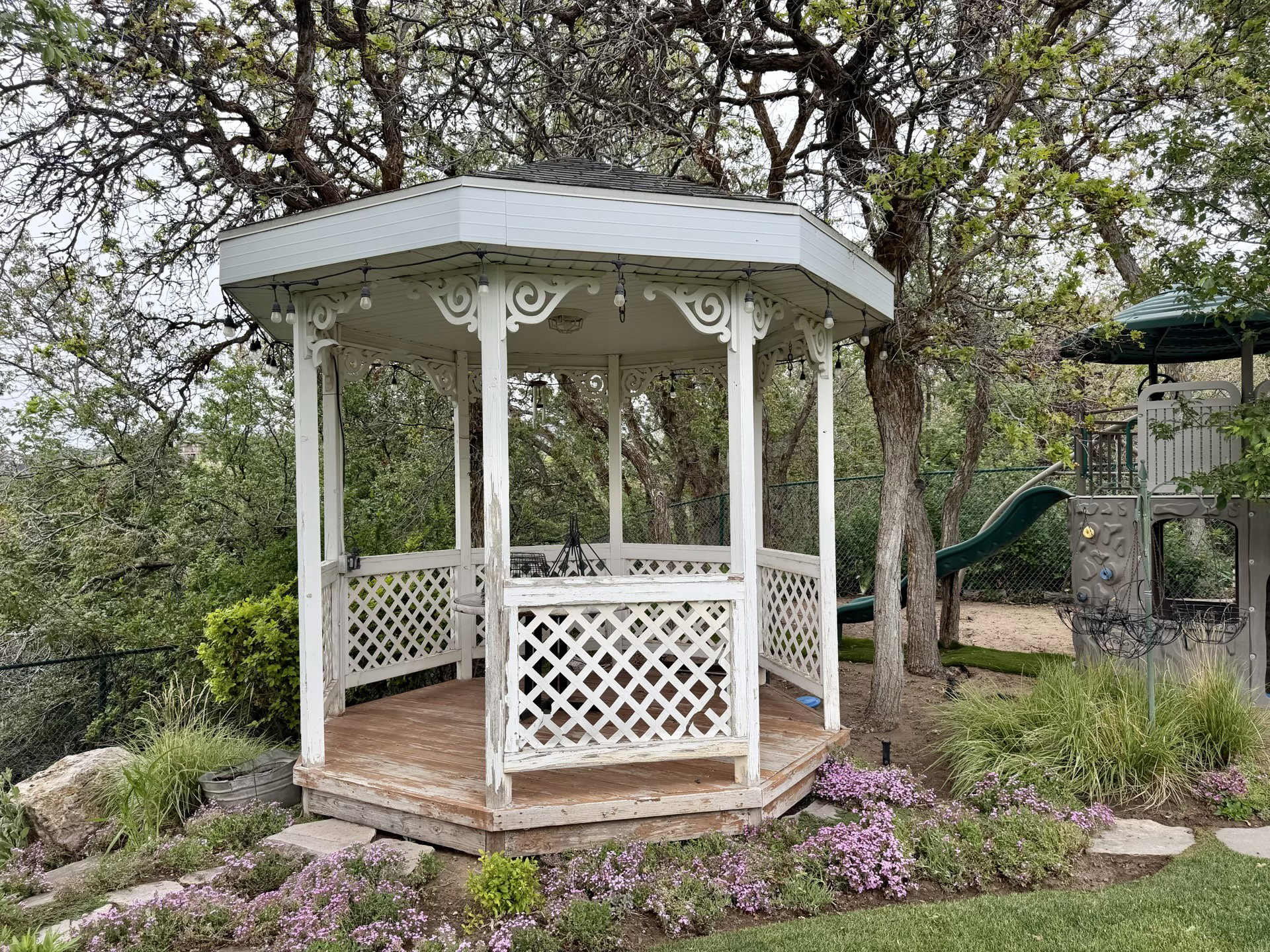 White octagonal gazebo with decorative lattice work, wood-finish flooring, and string lighting