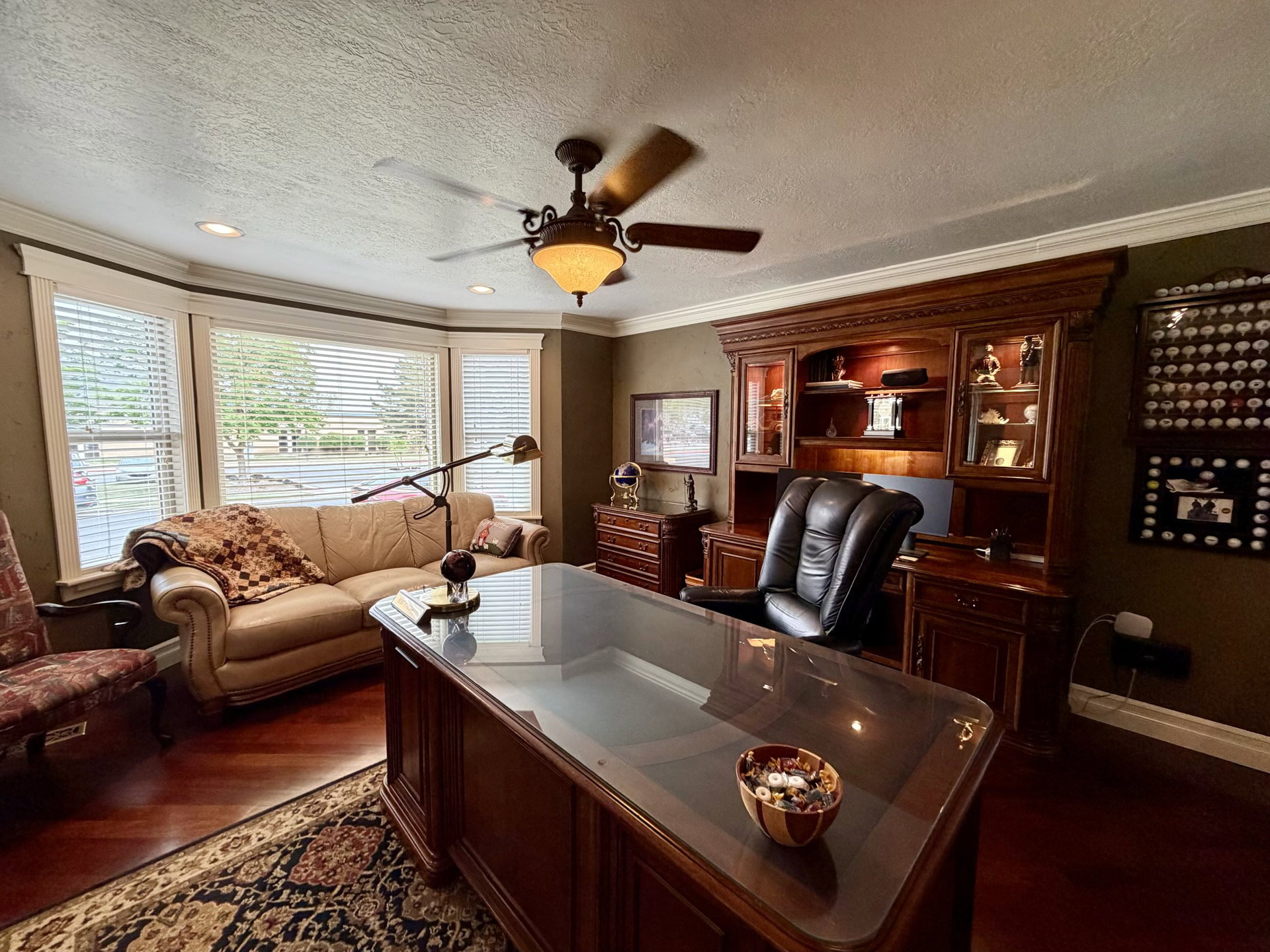 Dedicated room featuring rich wood-finish flooring, a prominent bay window with blinds, a built-in wood-finish cabinet with display lighting, and a ceiling fan with integrated lighting