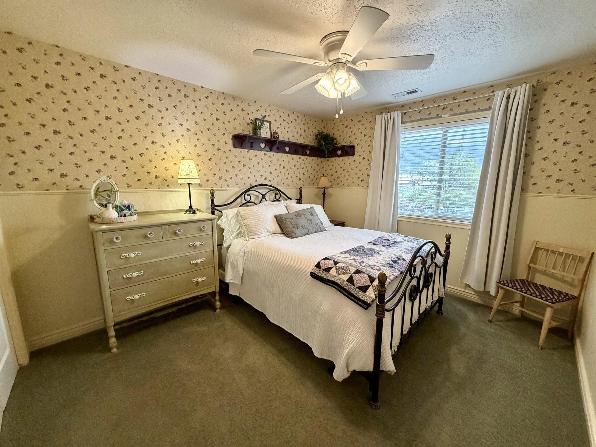 Carpeted room featuring a ceiling fan with integrated lighting, a window with blinds and drapes, and wainscoting with patterned wallpaper above