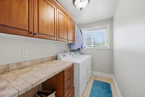 Dedicated laundry space featuring upper and lower wood cabinetry, a tiled countertop, and a window with blinds