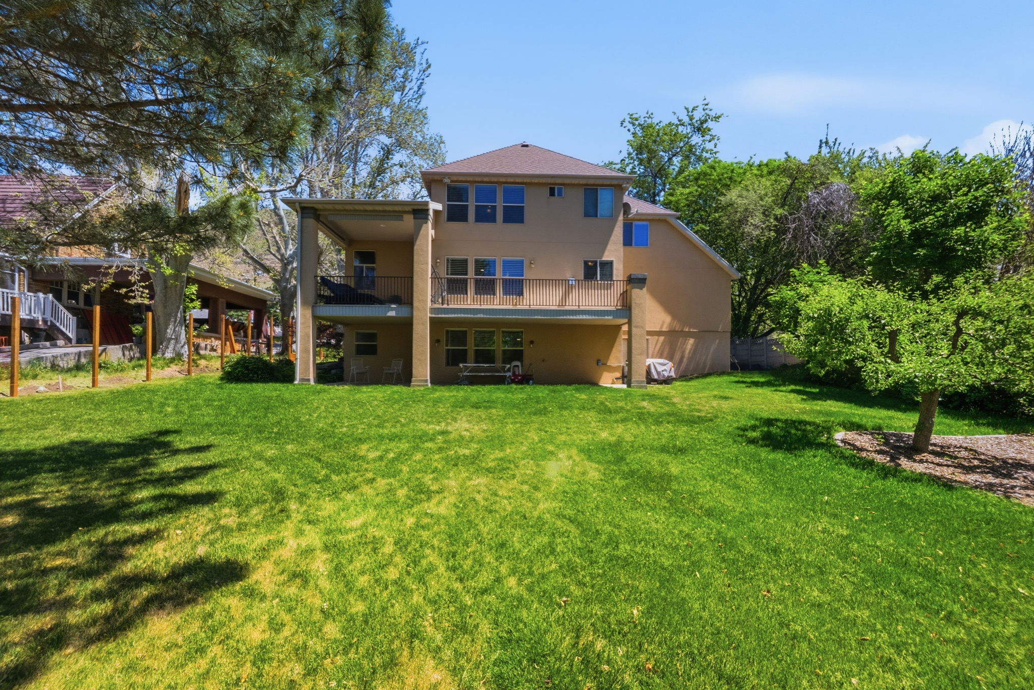 Expansive grassy yard featuring a multi-story residence with a covered patio, upper-level balcony, multiple large windows, and mature perimeter trees