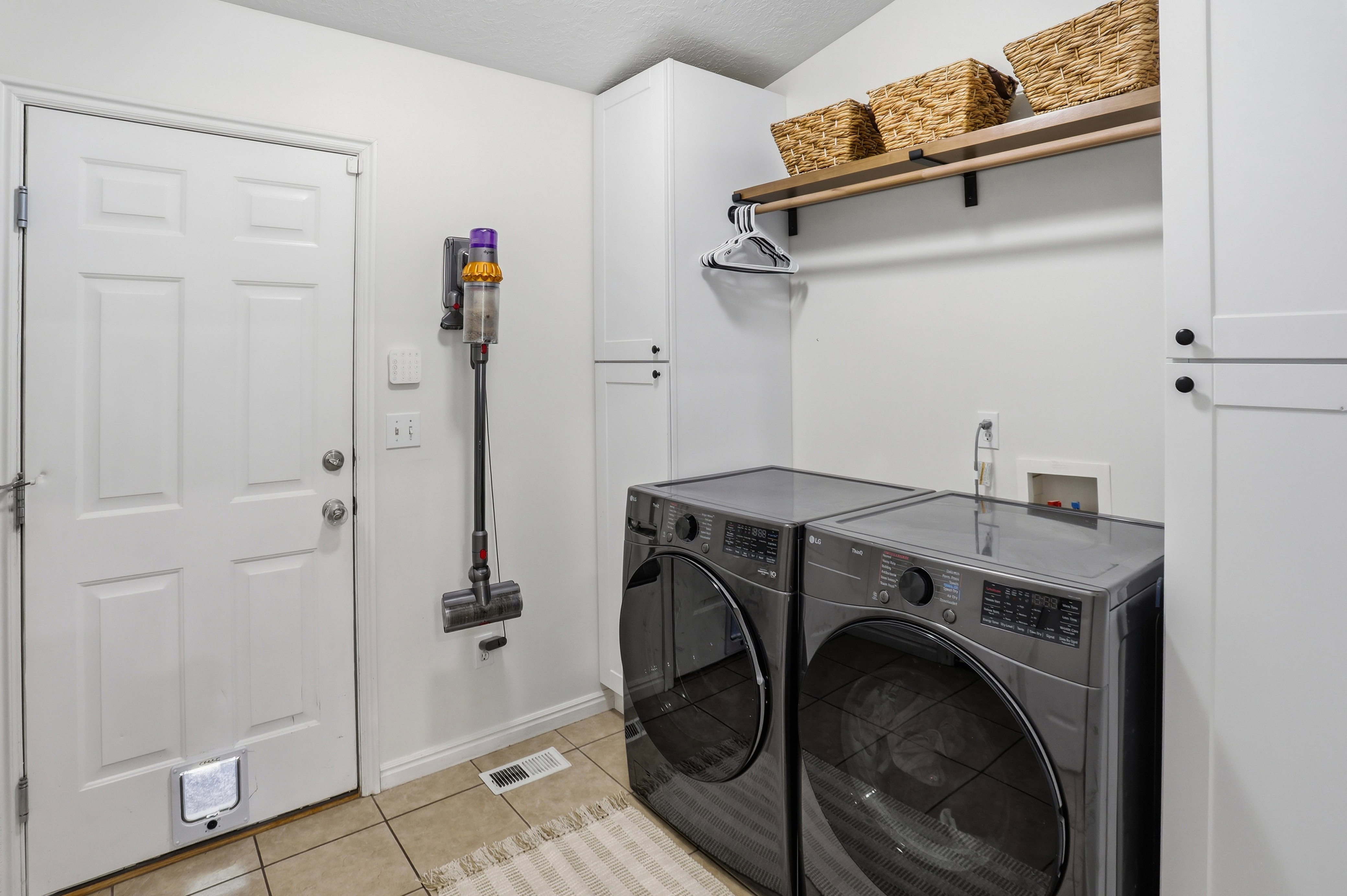 Dedicated laundry area featuring modern front-loading appliances, white cabinetry, a wood shelf with integrated hanging rod, and tile flooring
