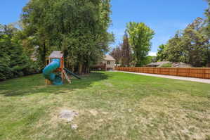 Expansive green space featuring a wooden play structure with a slide, mature trees, and a privacy fence
