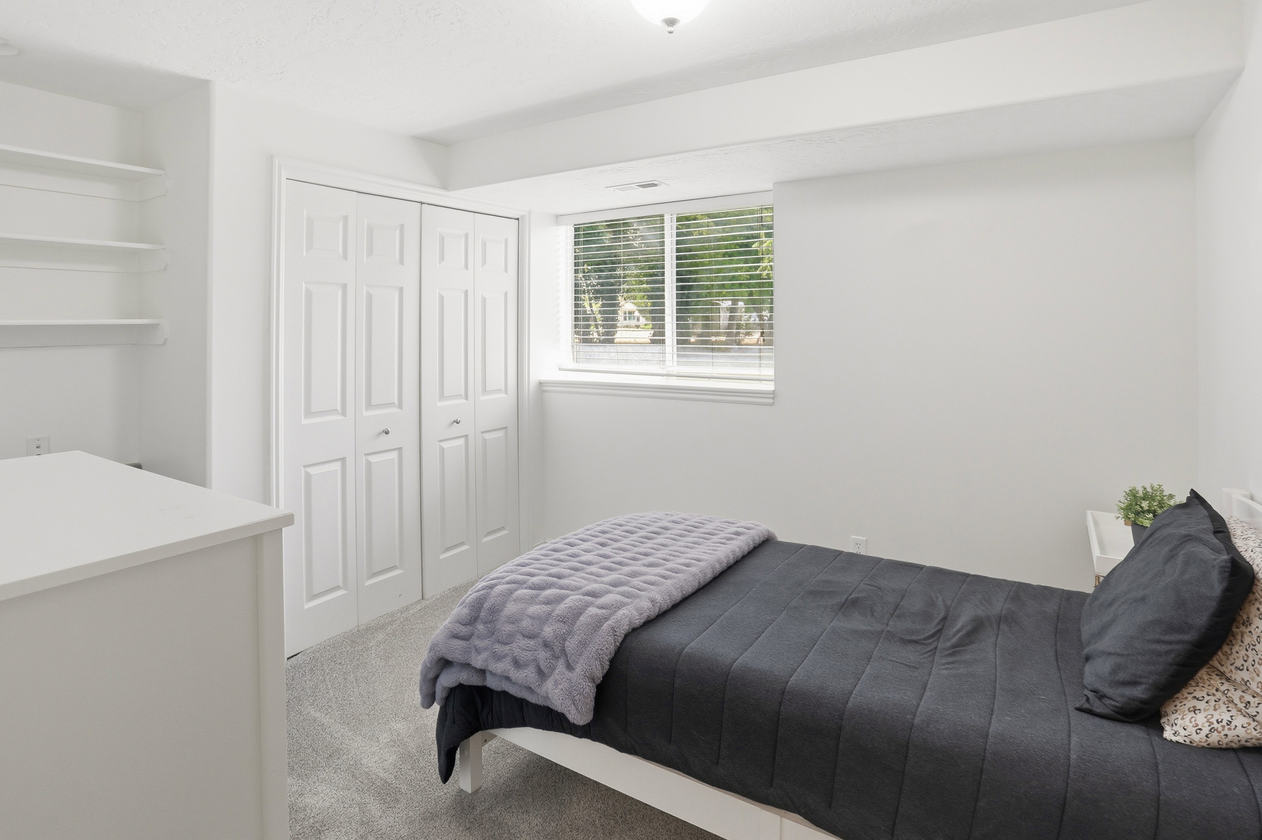 Bright interior space featuring a window with horizontal blinds, built-in shelving, bi-fold closet doors, and light grey carpet flooring