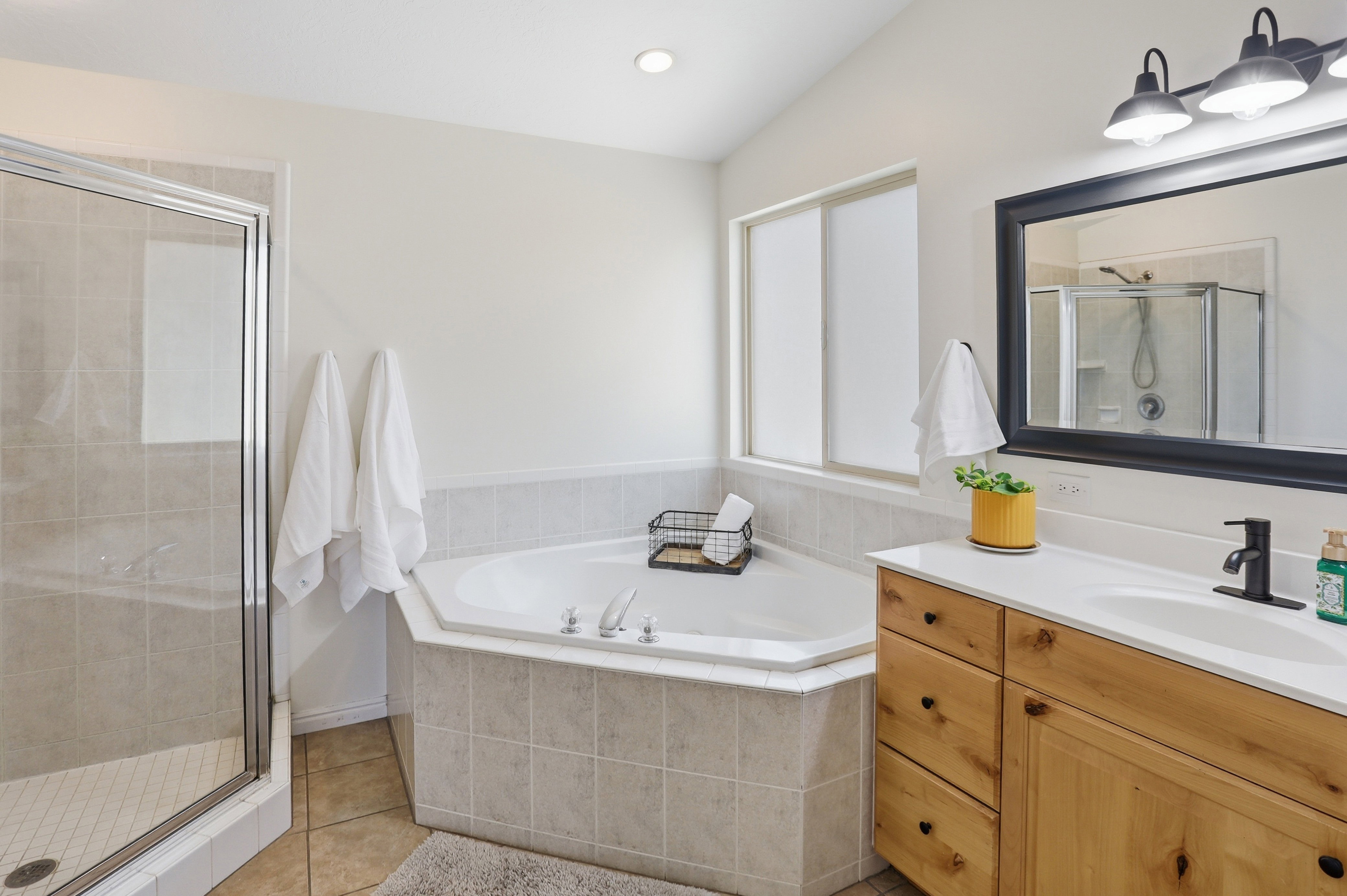 Master bathroom featuring a corner soaking tub with tile surround, a glass-enclosed shower with tile walls, a vanity with wood cabinetry and a white countertop, and a large mirror with a dark frame
