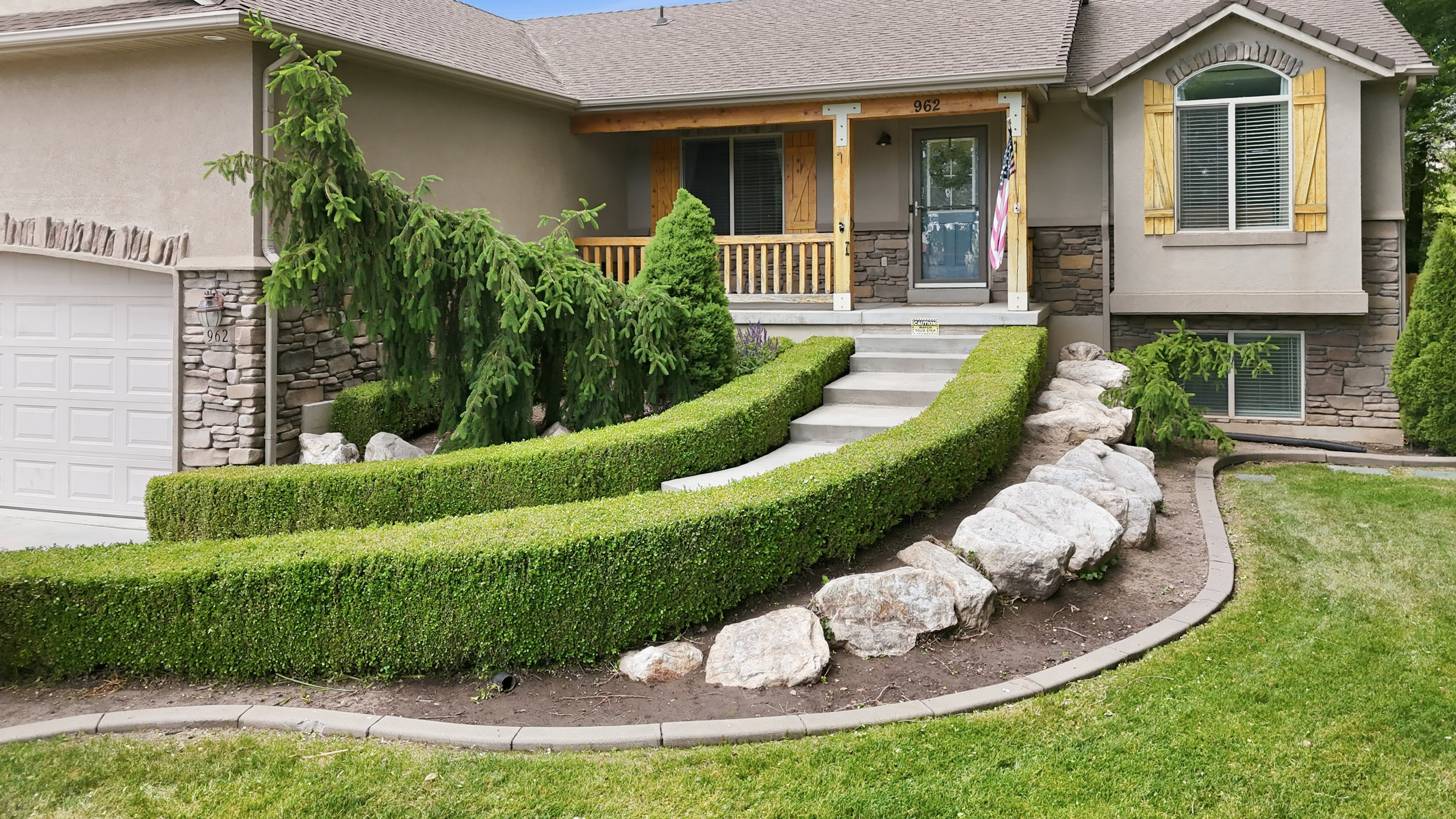 Front exterior featuring varied landscaping with manicured hedges, decorative boulders, and mature plantings