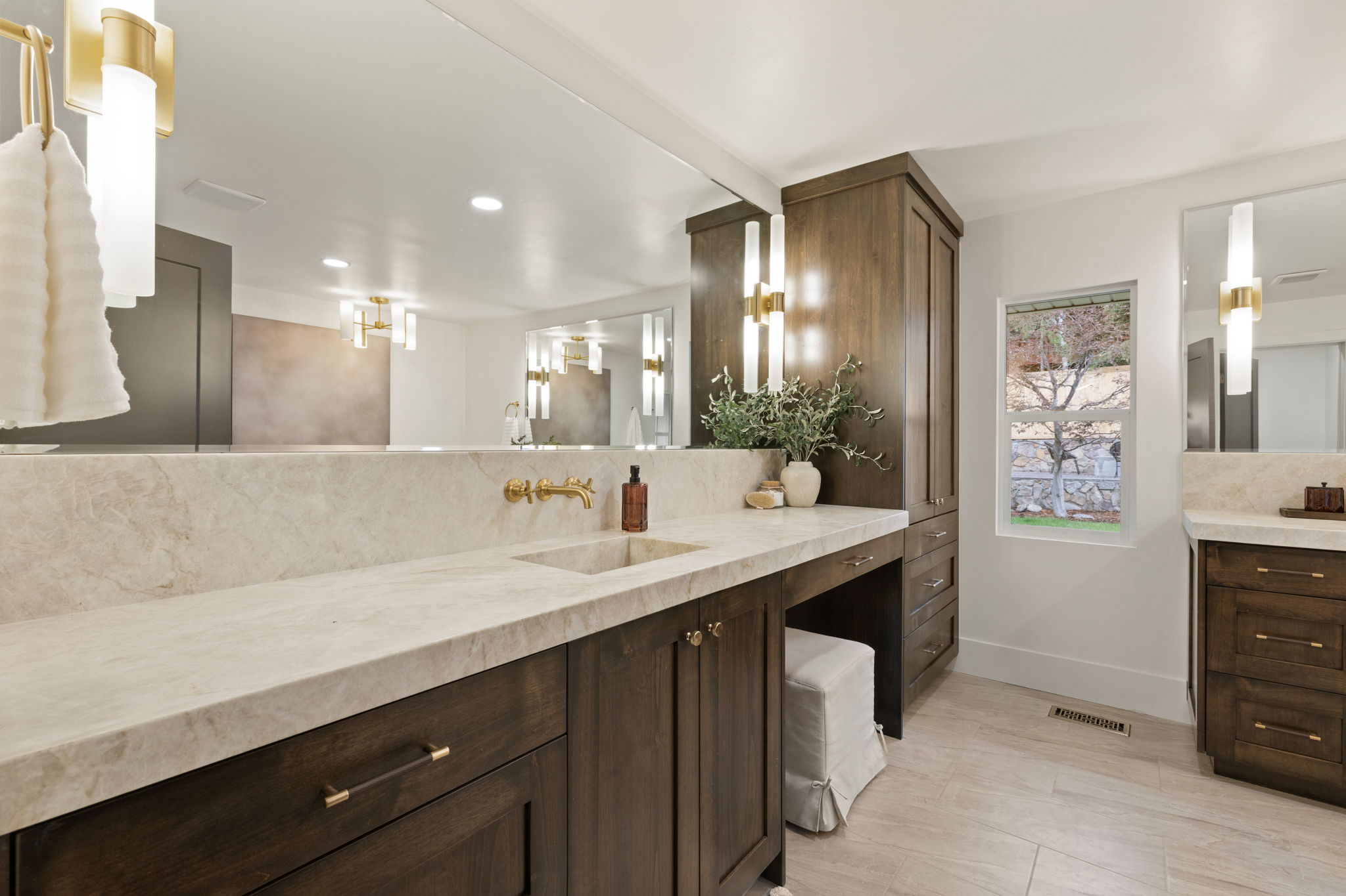 Spacious vanity area featuring light-toned countertops and backsplash, under-mount sink, wall-mounted brass faucet, and dark wood cabinetry with brass hardware