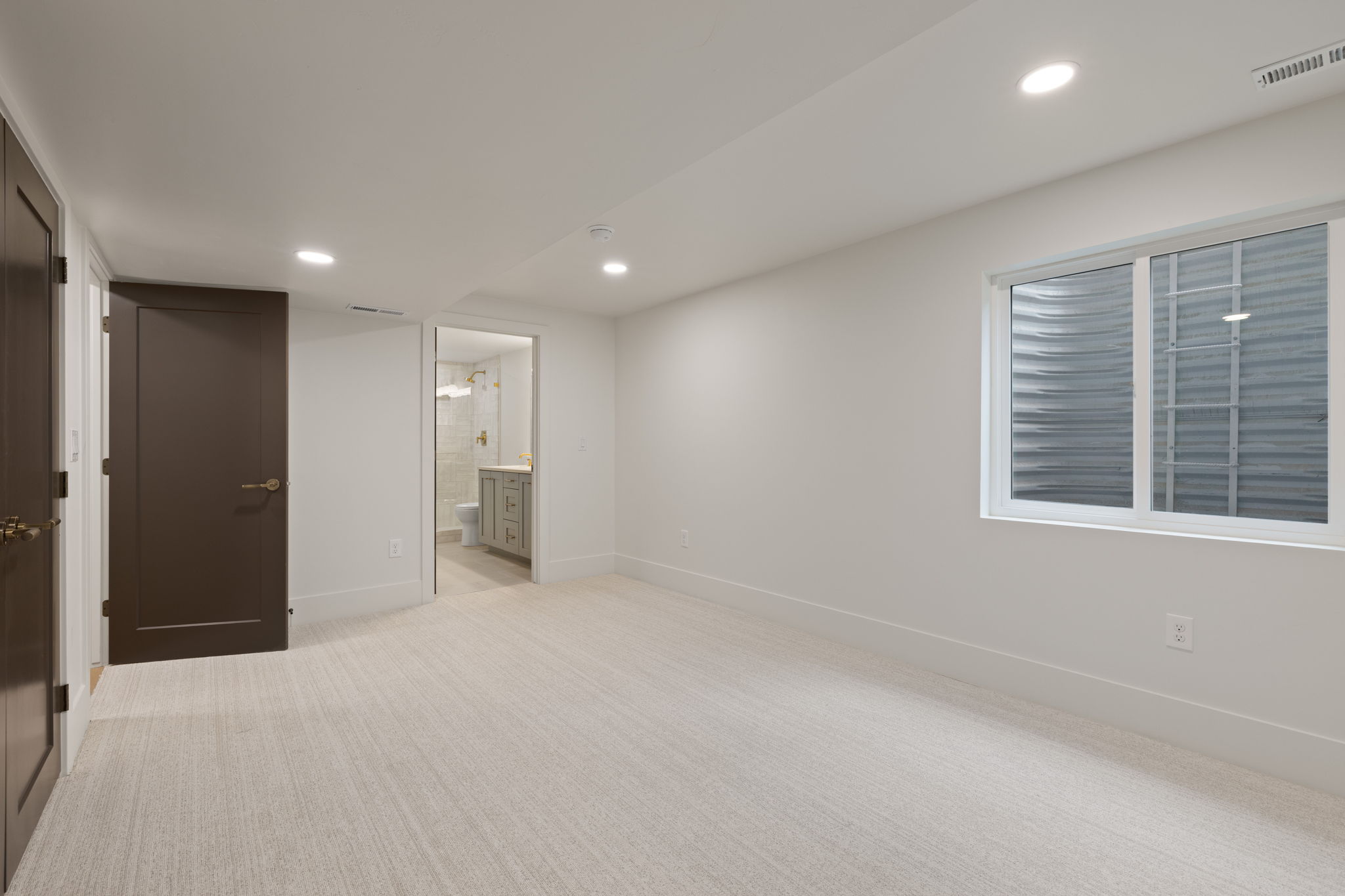Spacious room featuring light-toned carpet flooring, a large window with privacy blinds, recessed lighting, and a dark wood-finish interior door