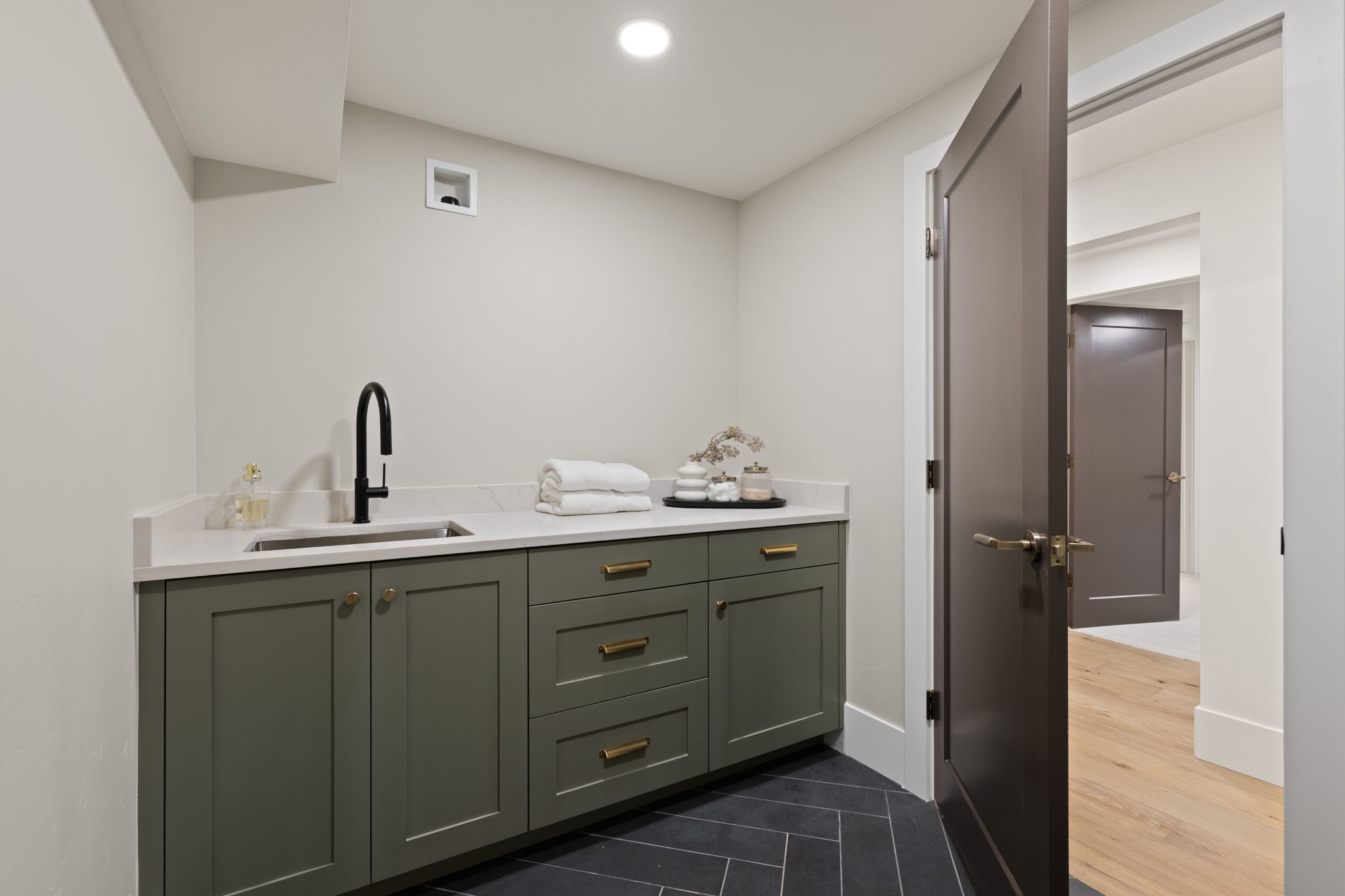 Laundry room featuring a stainless steel sink with a matte black faucet, light quartz countertop, and green shaker-style cabinetry with brass hardware