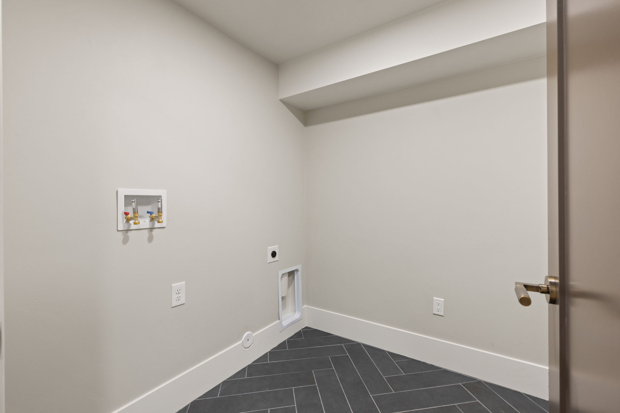 Dedicated laundry area featuring modern grey herringbone-patterned tile flooring, fresh light grey wall paint, white baseboards, and essential utility connections
