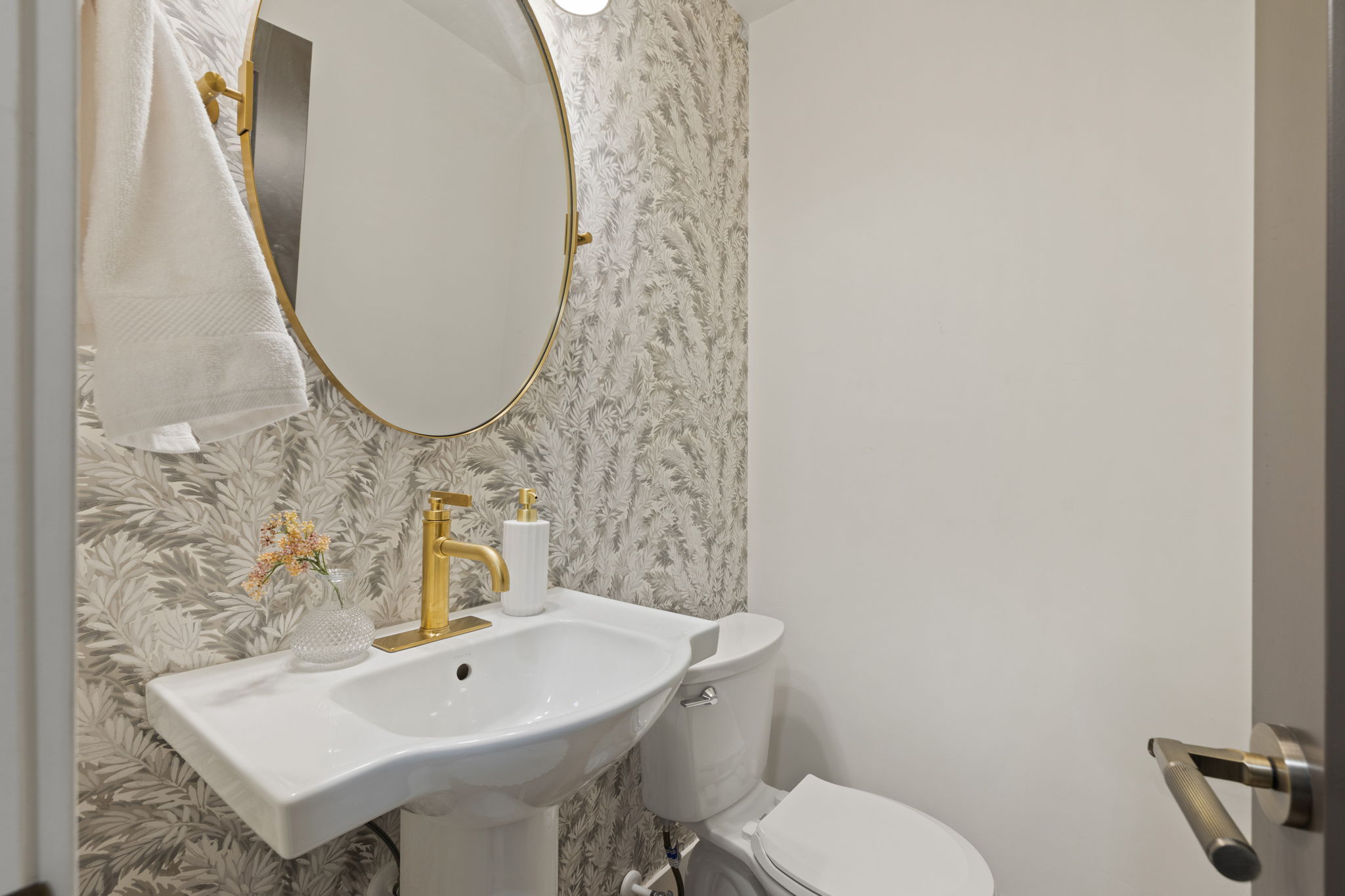 Pedestal sink with gold-finish faucet, oval mirror, and decorative wall covering