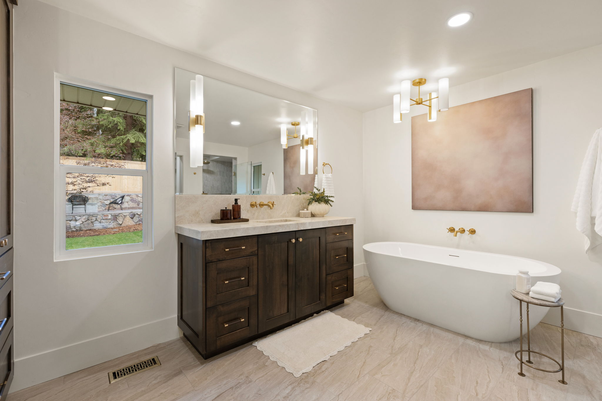 Spacious bathroom featuring a freestanding soaking tub, dual-sink vanity with dark wood cabinetry and light-colored countertop, large wall-mounted mirror, and stylish brass lighting fixtures