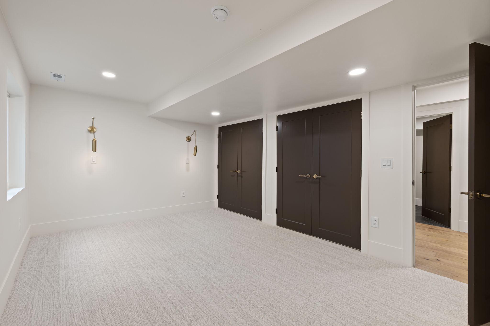 Spacious room featuring light-toned carpeting, recessed ceiling lighting, two sets of dark panel doors with brass hardware, and two decorative wall sconces