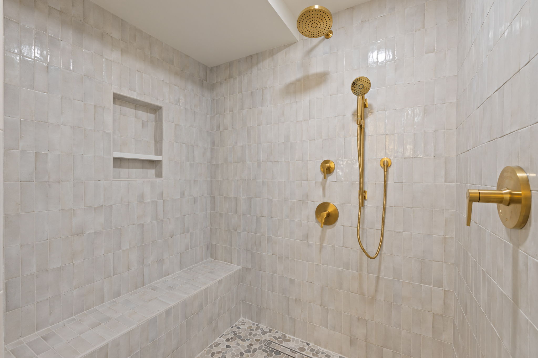 Walk-in shower featuring light-toned square wall tiles, a built-in shower bench, and gold-finish fixtures including a rain showerhead and handheld sprayer